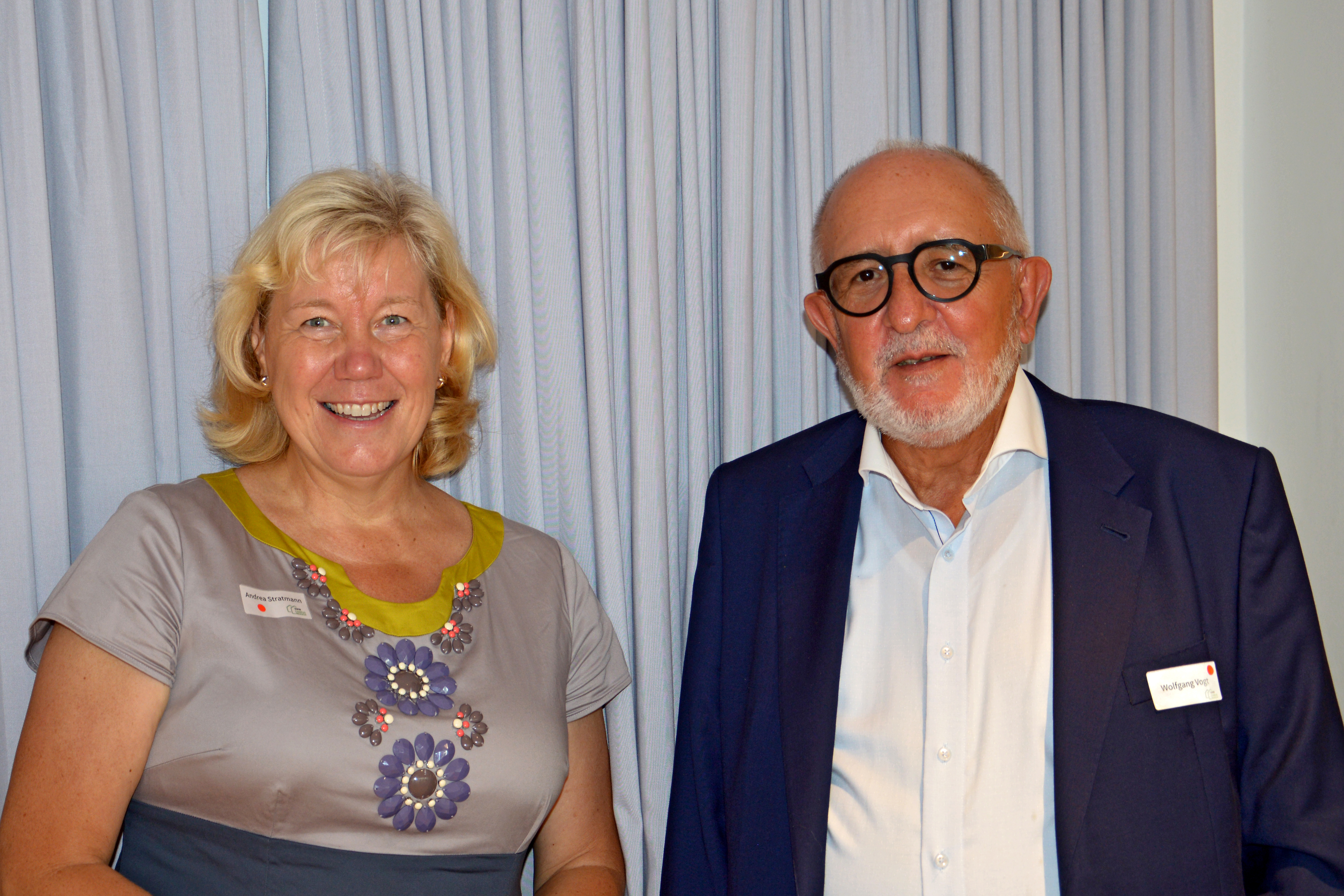 Porträtaufnahme einer freundlich lachenden Frau mit blonden mittellangen Haaren in einem grauen Kleid mit lila Blumen und einem lächelnden Herrn mit dunkler Brille und hellem Bart in einem blauen Anzug.