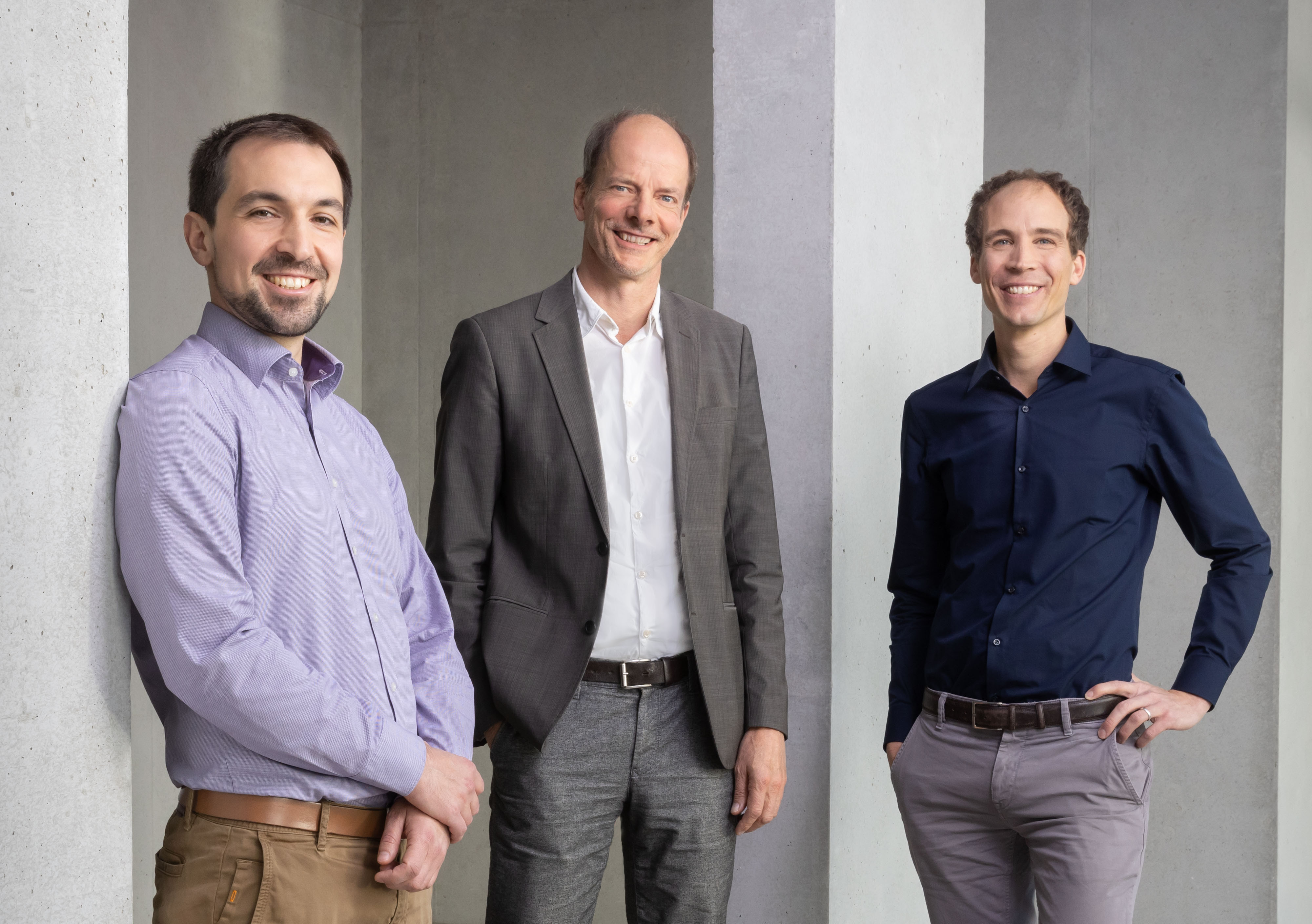 Three young men in shirts stand in front of a concrete wall and smile friendly into the camera.