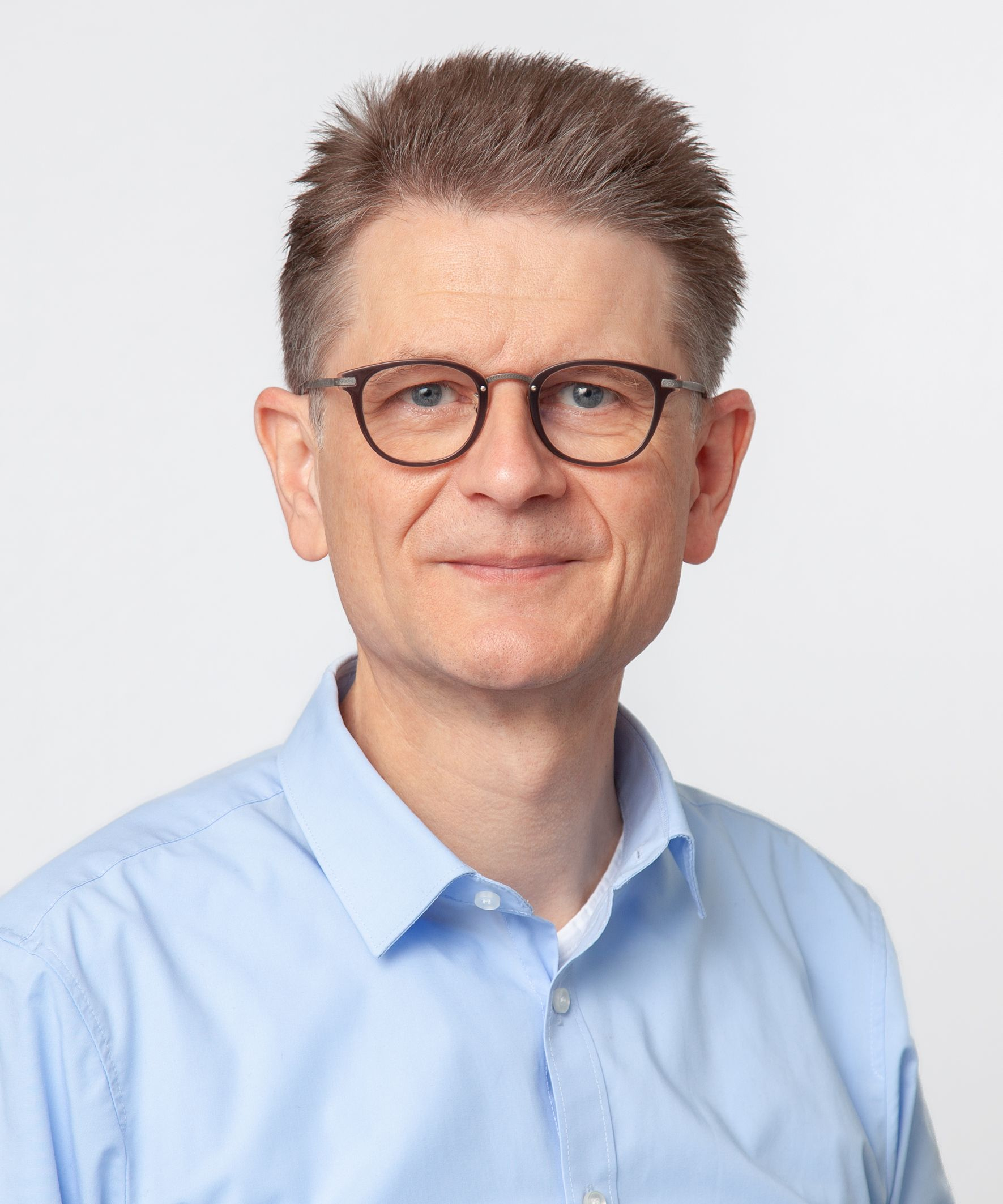 The photo shows a man with a blue shirt and glasses looking friendly into the camera.