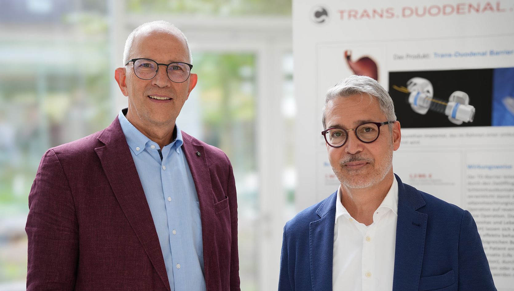 Zwei Männer im Anzug mit Brille und kurzen grauen Haaren stehen vor einem Poster, das den Barrier erklärt.