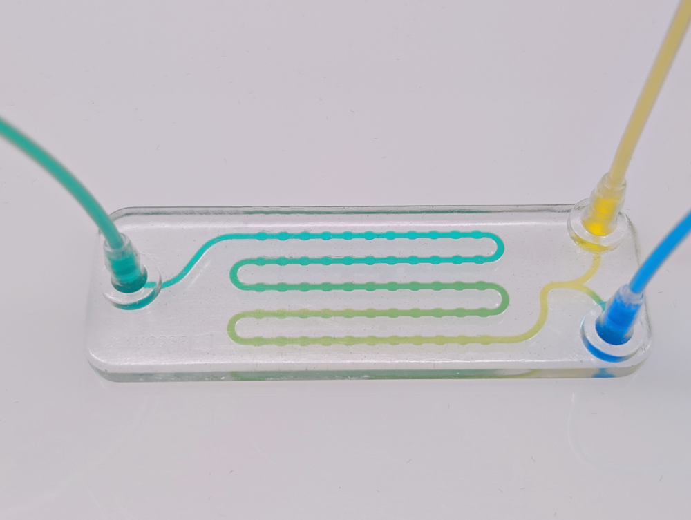 Rectangular glass chip with three tubes at one end, through which yellow, green and blue liquids flow into the chip.