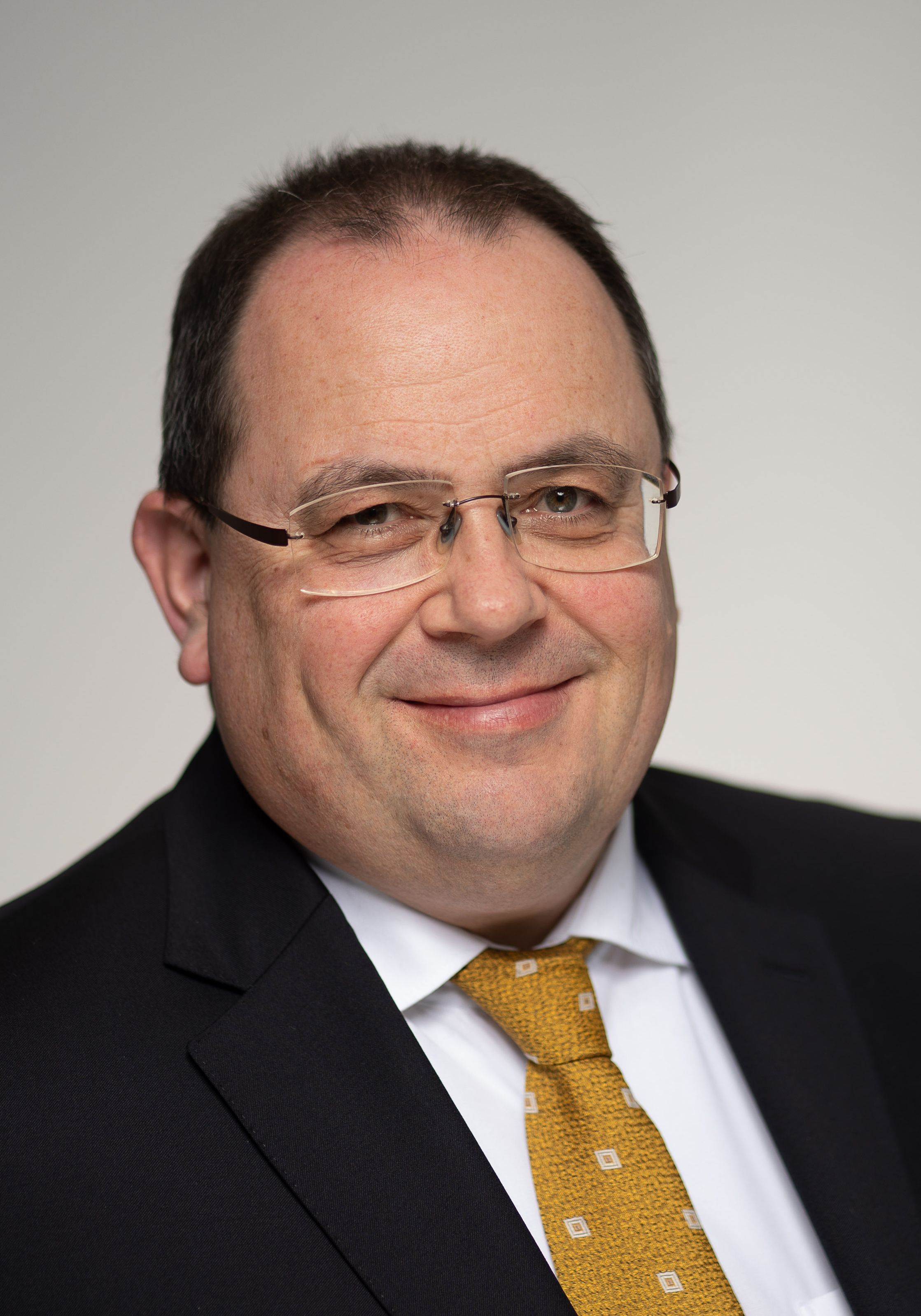 Portrait photo of a friendly smiling man in a black jacket with mustard-coloured tie and rimless glasses