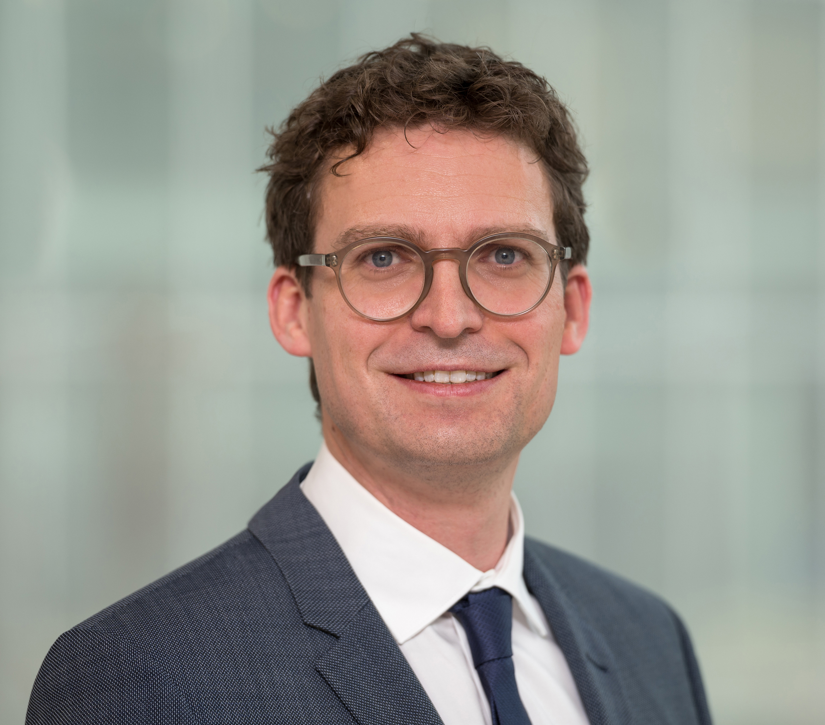 Portrait of a friendly smiling man in a suit with dark blond hair and glasses.