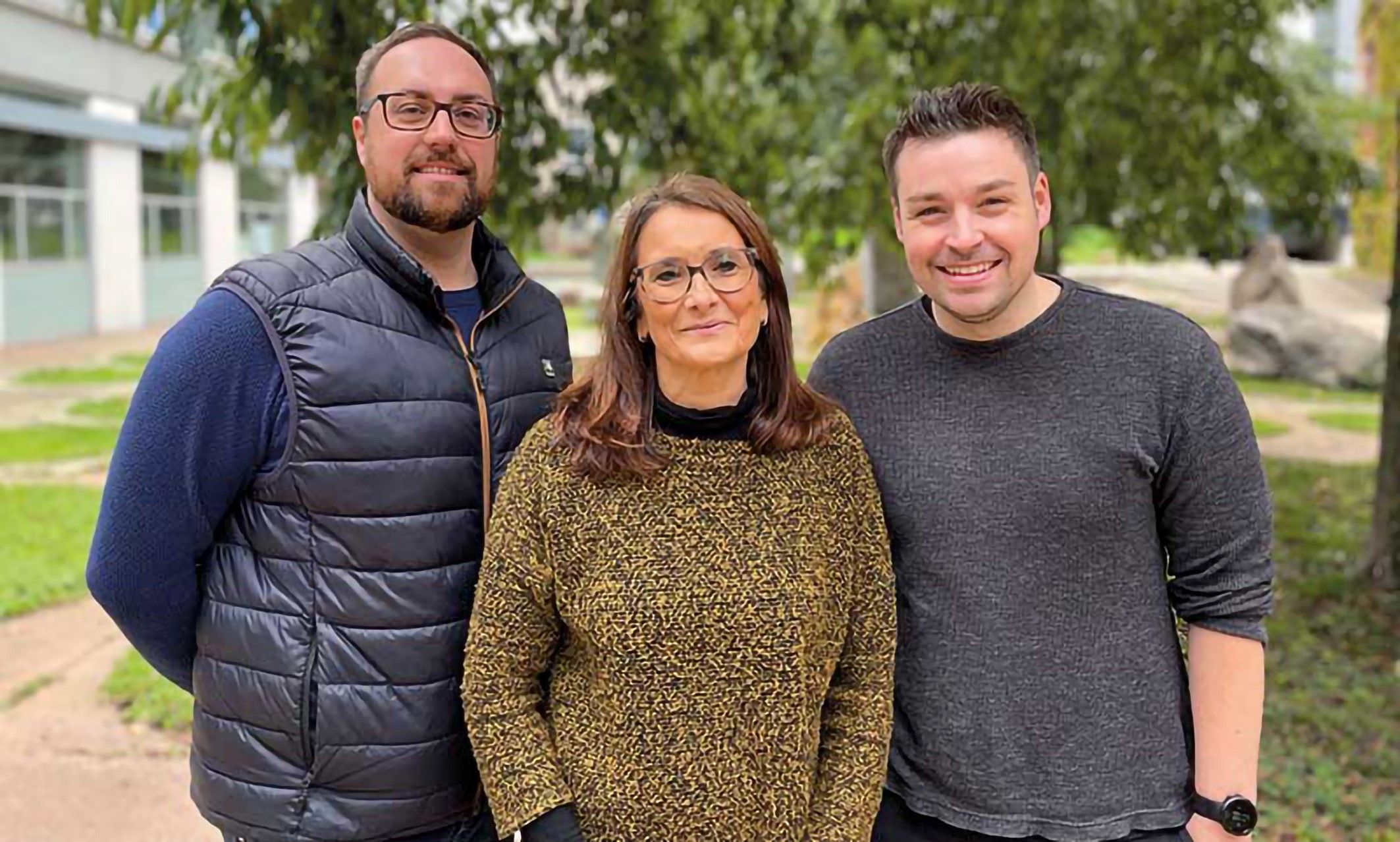 The photo shows two men and in the middle a woman standing in front of a tree and smiling at the camera.