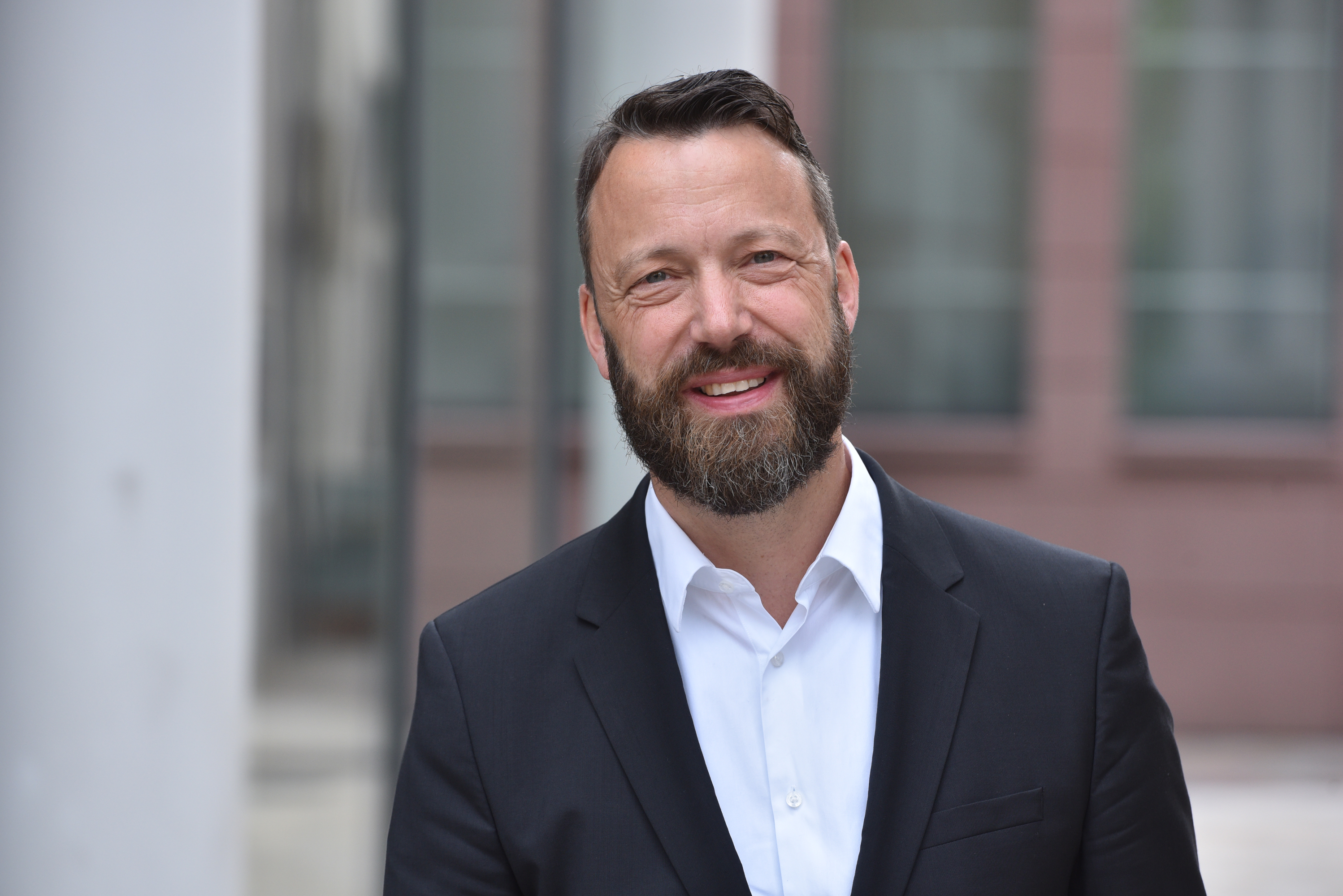 Portrait photo of friendly looking man with beard wearing white shirt and dark gray jacket.