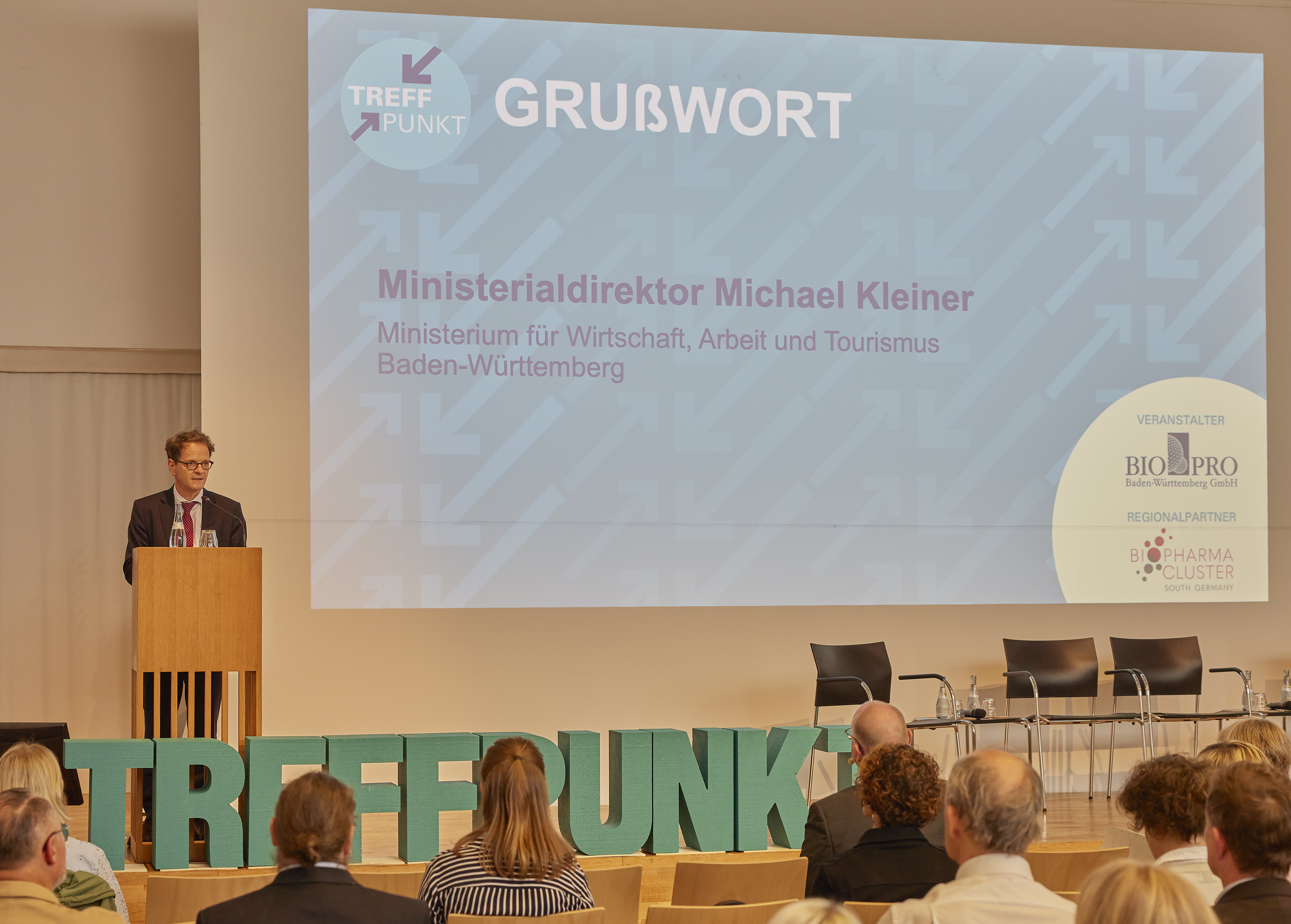 Eine Präsentation in einem Konferenzraum, bei der auf der Leinwand "GRUßWORT" und der Name "Ministerialdirektor Michael Kleiner, Ministerium für Wirtschaft, Arbeit und Tourismus Baden-Württemberg" zu lesen sind, während unten im Vordergrund grüne Buchstaben "Treffpunkt" zu sehen sind.