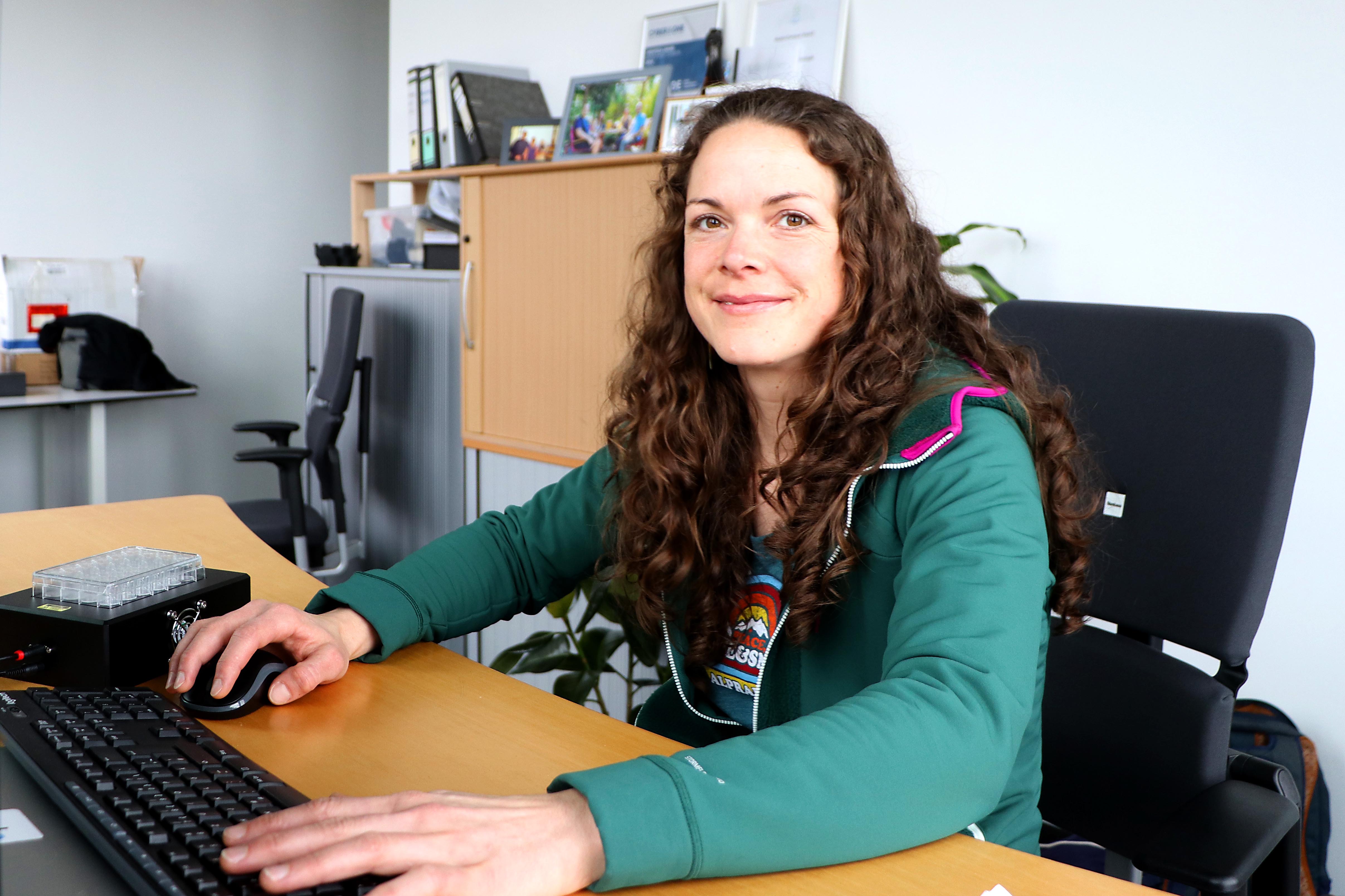 A young woman at a desk with long brown curls can be seen looking friendly into the camera.