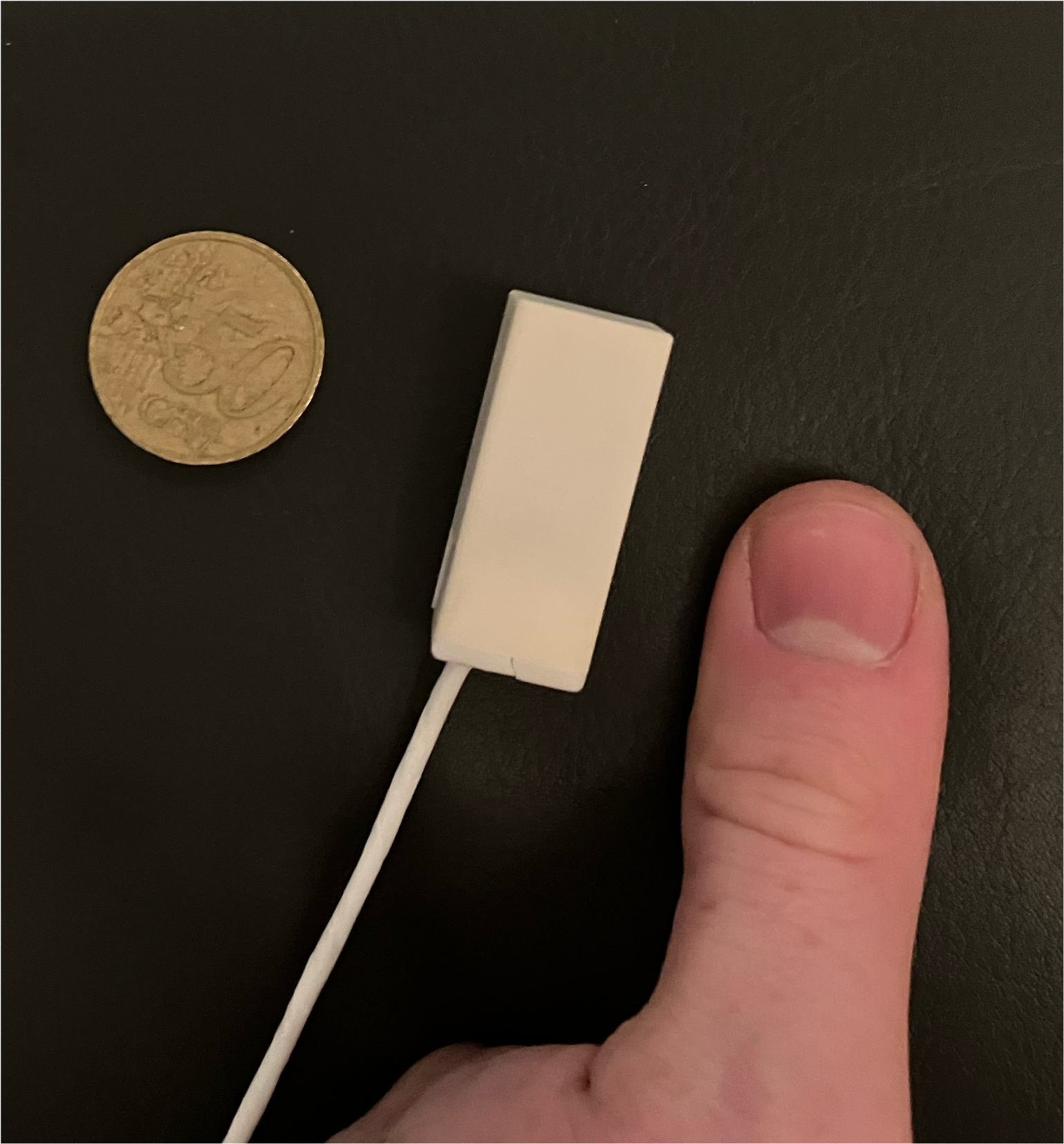 heller rechteckiger Sensor, links davon eine 50-Cent-Münze, die etwas breiter und etwas weniger lang ist. Rechts davon ein Daumen, der etwa doppelt so lang, aber etwas breiter ist.