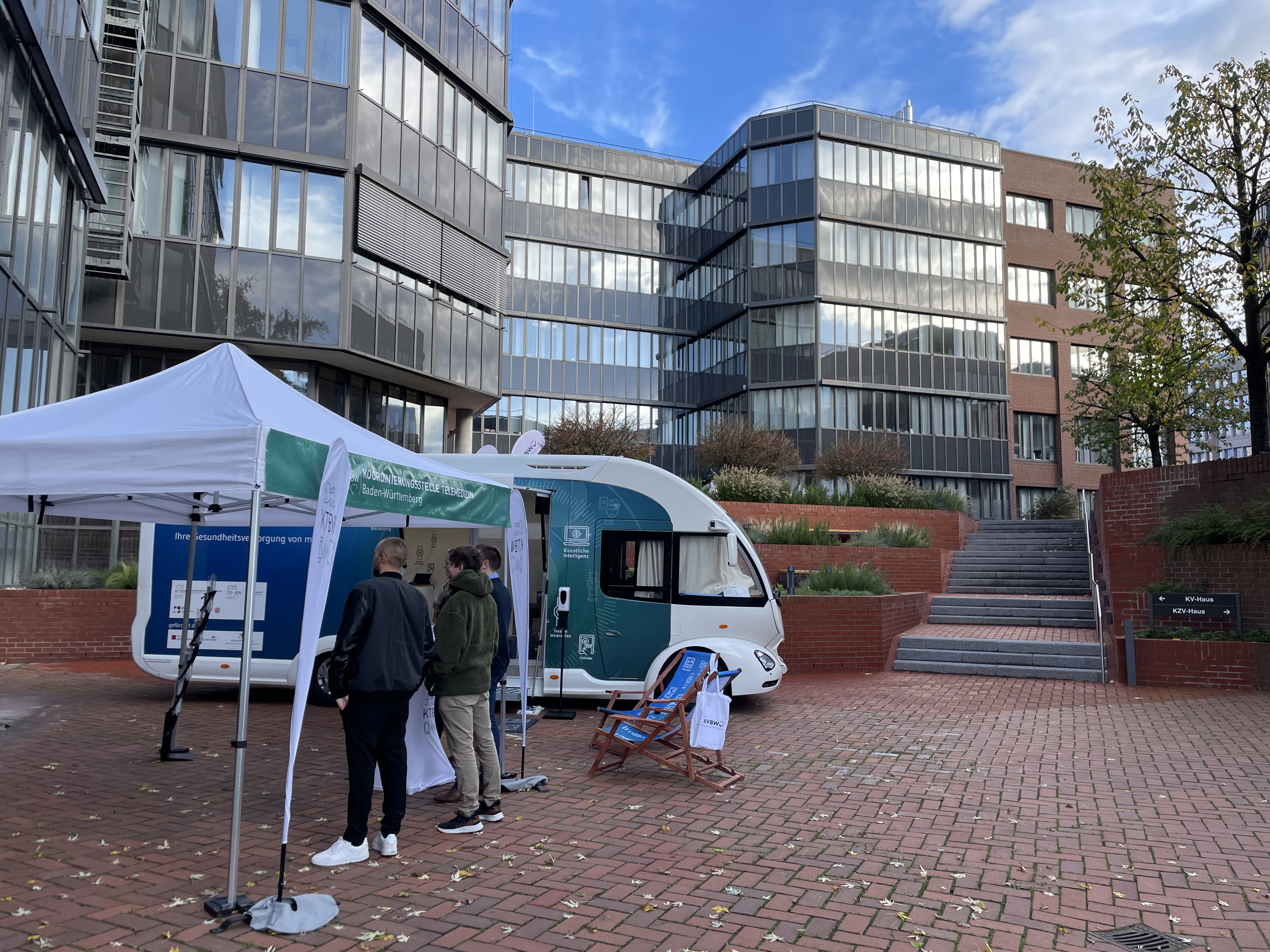 Der Truck steht mit dem Pavillon vor einem hohen Gebäude. Unter dem Pavillon stehen mehrere Menschen.