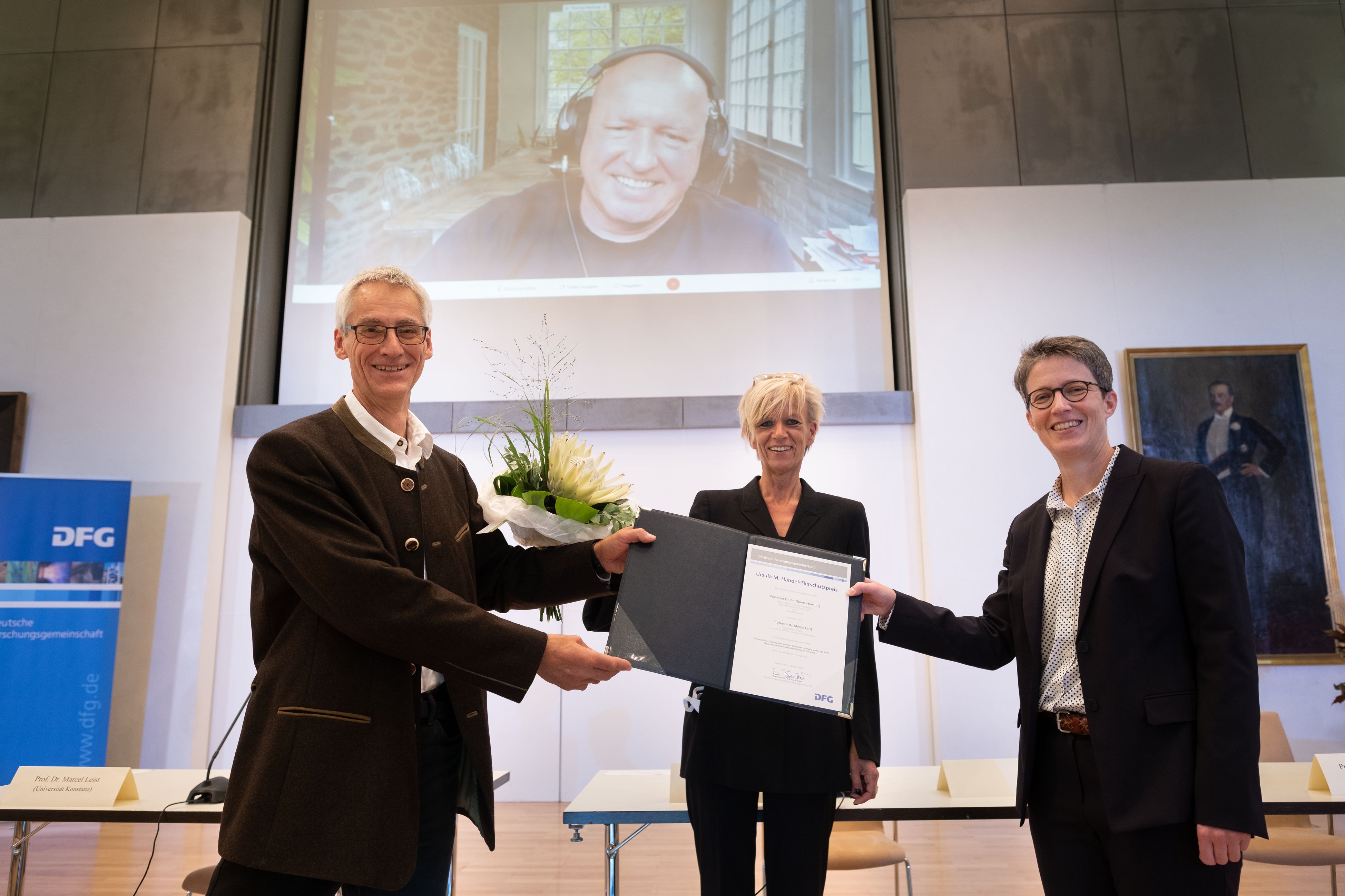 Prof. Marcel Leist und die DFG-Vizepräsidentin Prof. Dr. Britta Siegmund halten die Urkunde über den Ursula M. Händel-Tierschutzpreis der Deutschen Forschungsgemeinschaft (DFG) in der Hand. Ebenfalls im Bild: Die Vorsitzenden der Senatskommission für tierexperimentelle Forschung Prof. Dr. Brigitte Vollmar. Im Hintergrund auf einer Leinwand zu sehen ist Prof. Thomas Hartung, der online zugeschaltet ist.