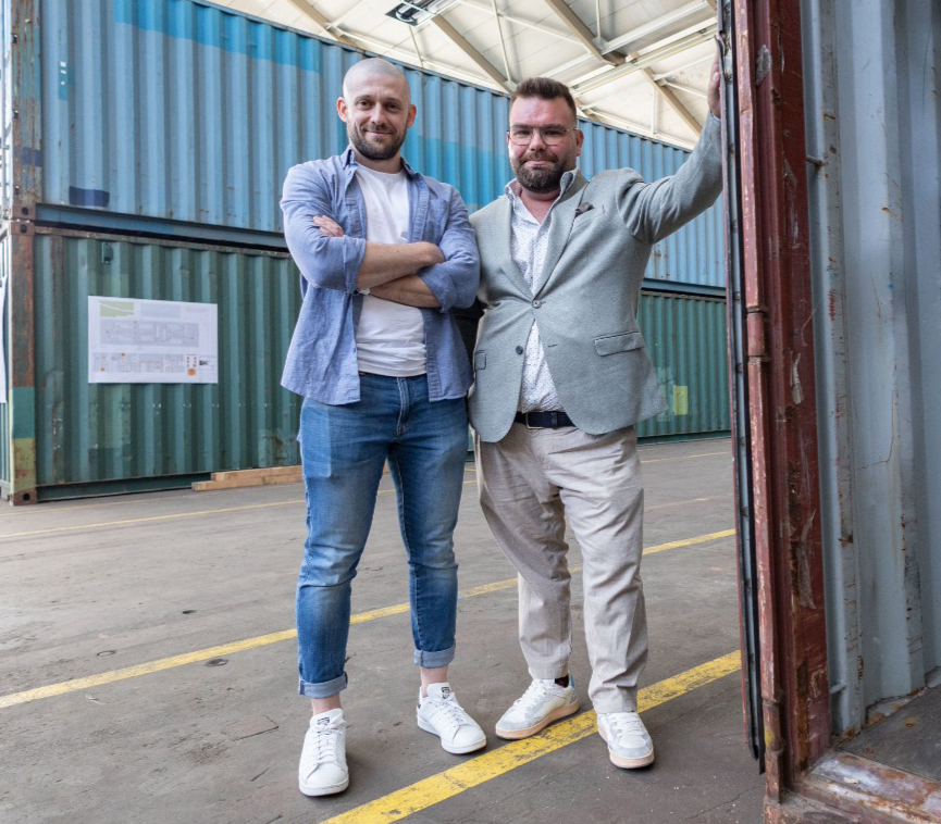 Zu sehen sind zwei junge Männer in legerer Kleidung in einer Industriehalle mit Containern im Hintergrund. Sie schauen beide direkt und lächelnd in die Kamera.