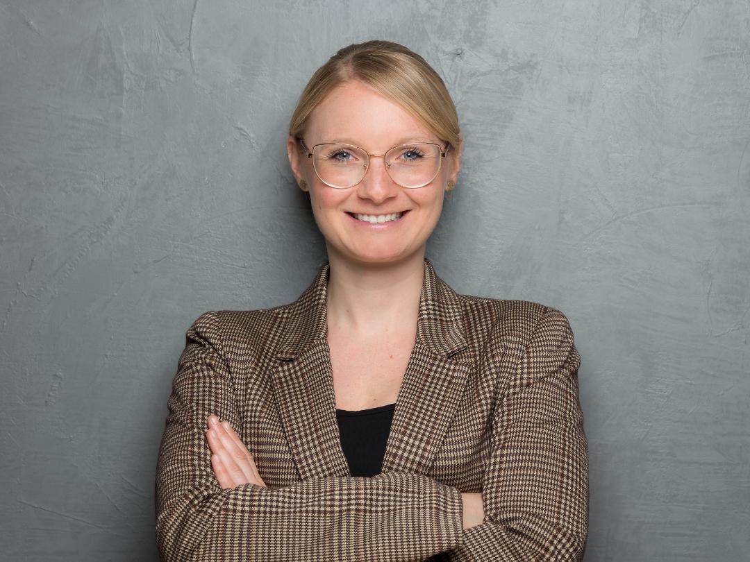 Zu sehen ist eine Frau mit Brille und kariertem Jackett, die mit verschränkten Armen freundlich in die Kamera lächelt.