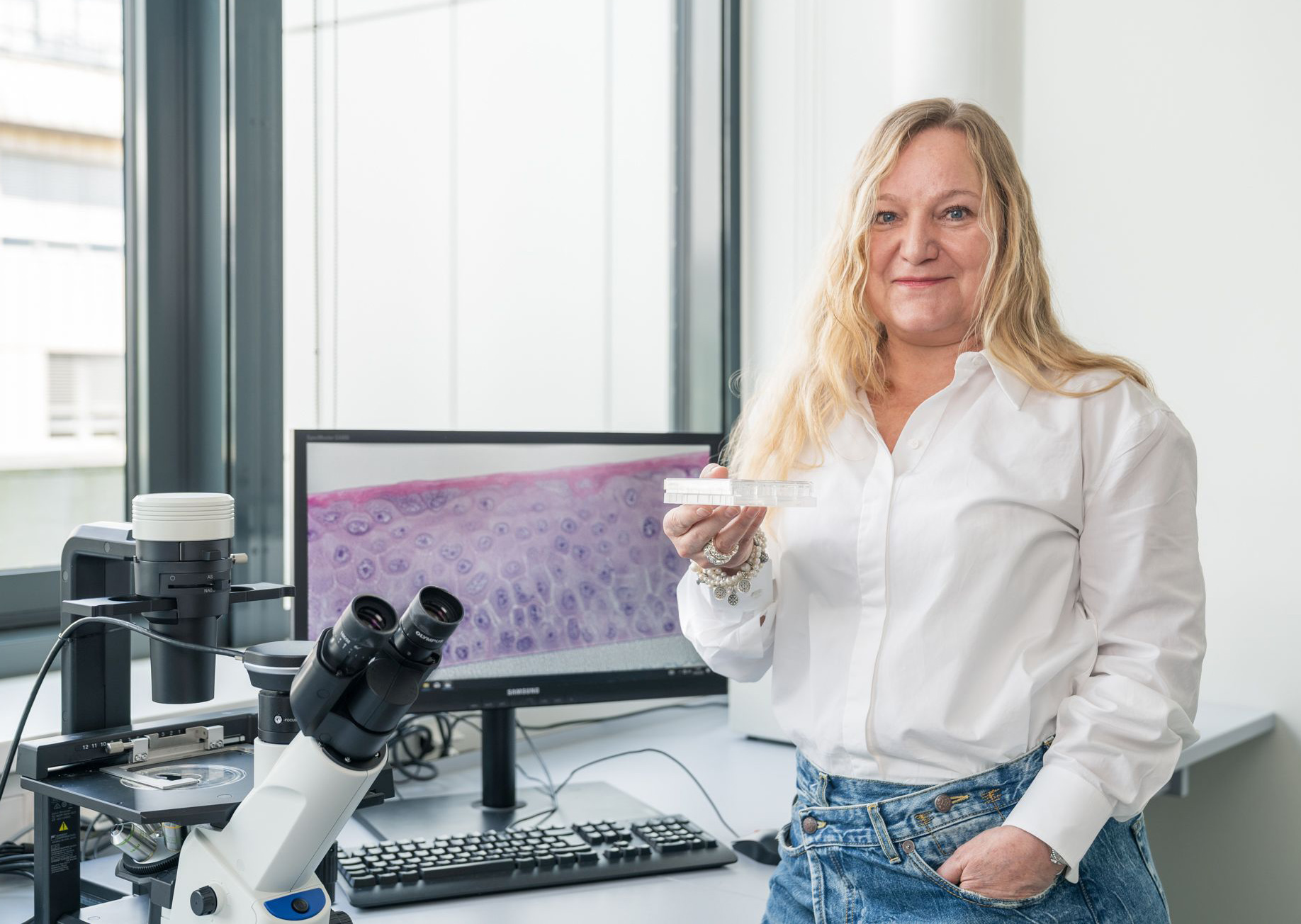 You can see a woman with long blonde hair standing in front of a PC screen and a microscope, holding a well plate in her hand. She smiles friendly into the camera.