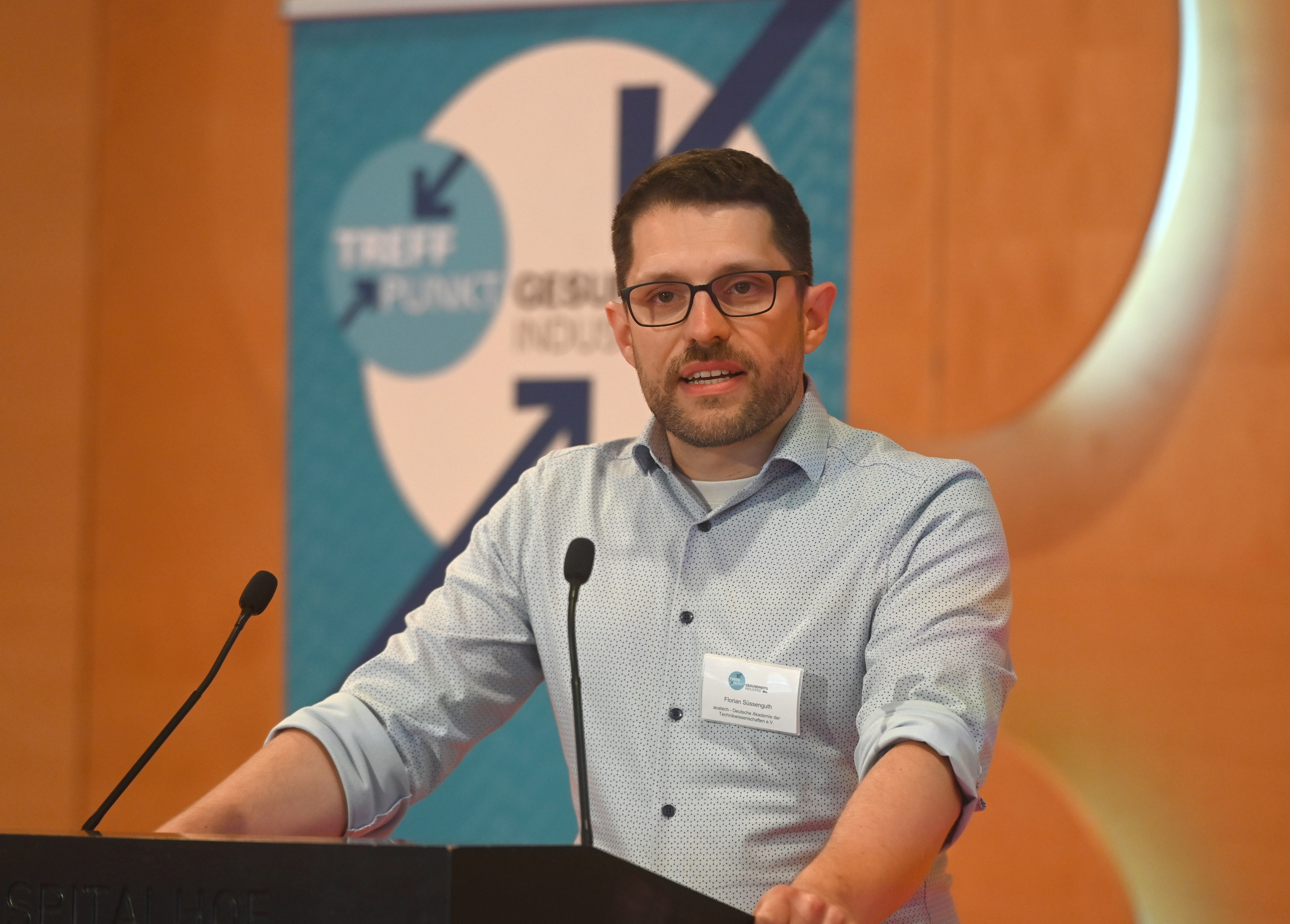 Ein dunkelhaariger Mann mit hellblauem Hemd und Brille vor einem Podium.