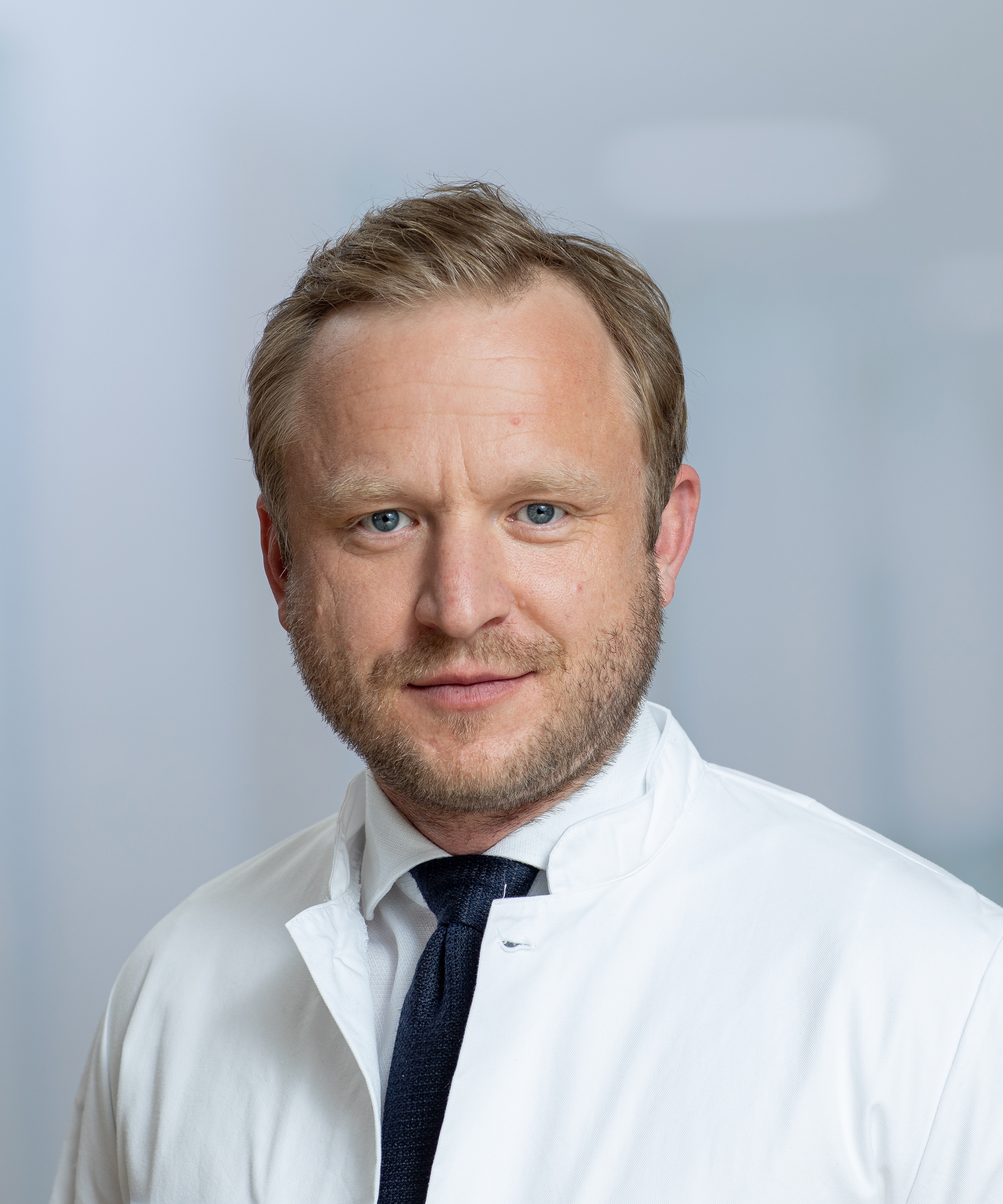 Portrait photograph of a man with medium blond short hair and three-day beard wearing a shirt and tie in a doctor's coat.