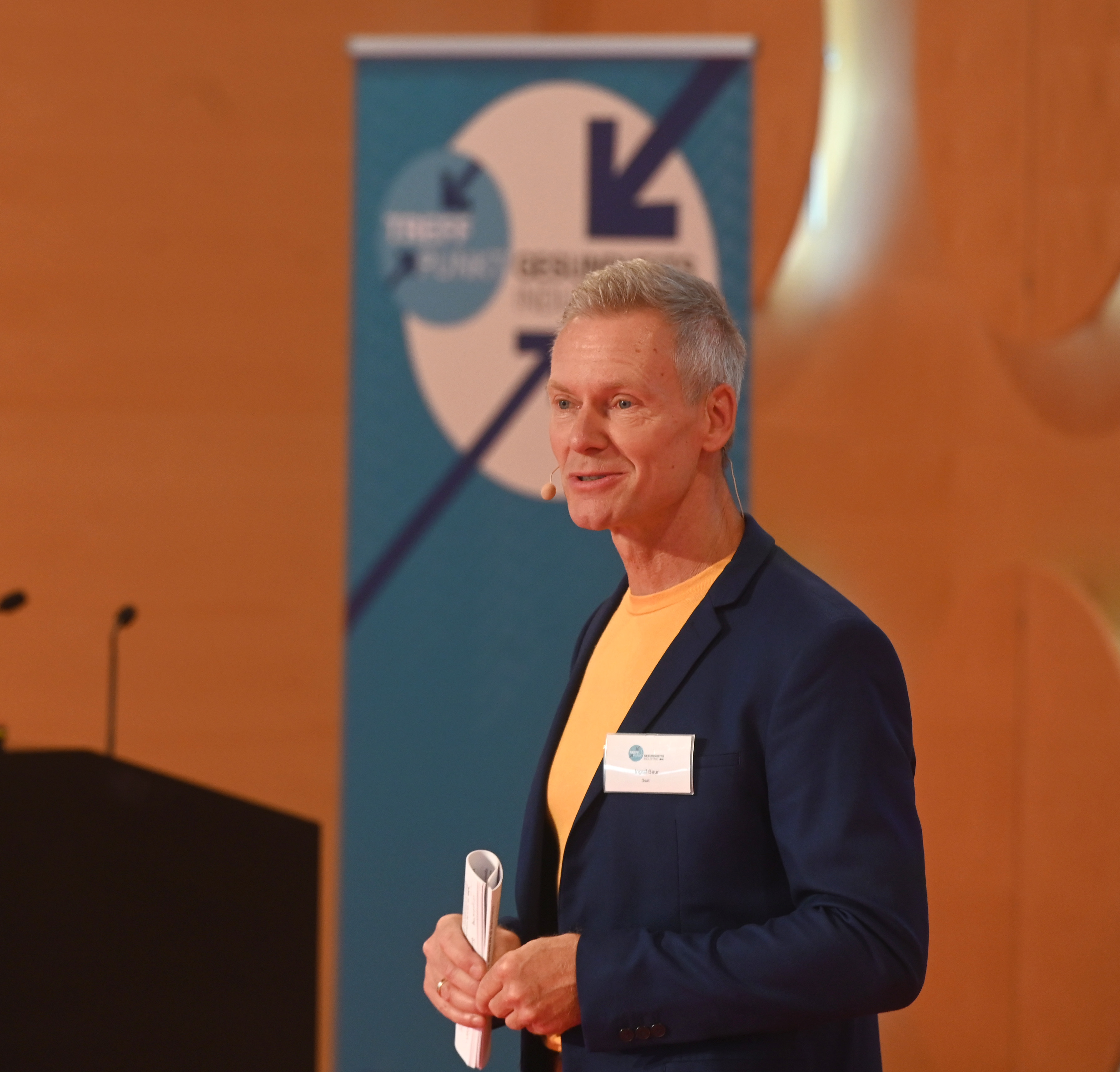 Ein hellblonder Mann mit gelbem T-Shirt und blauer Anzugsjacke auf einem Podium.