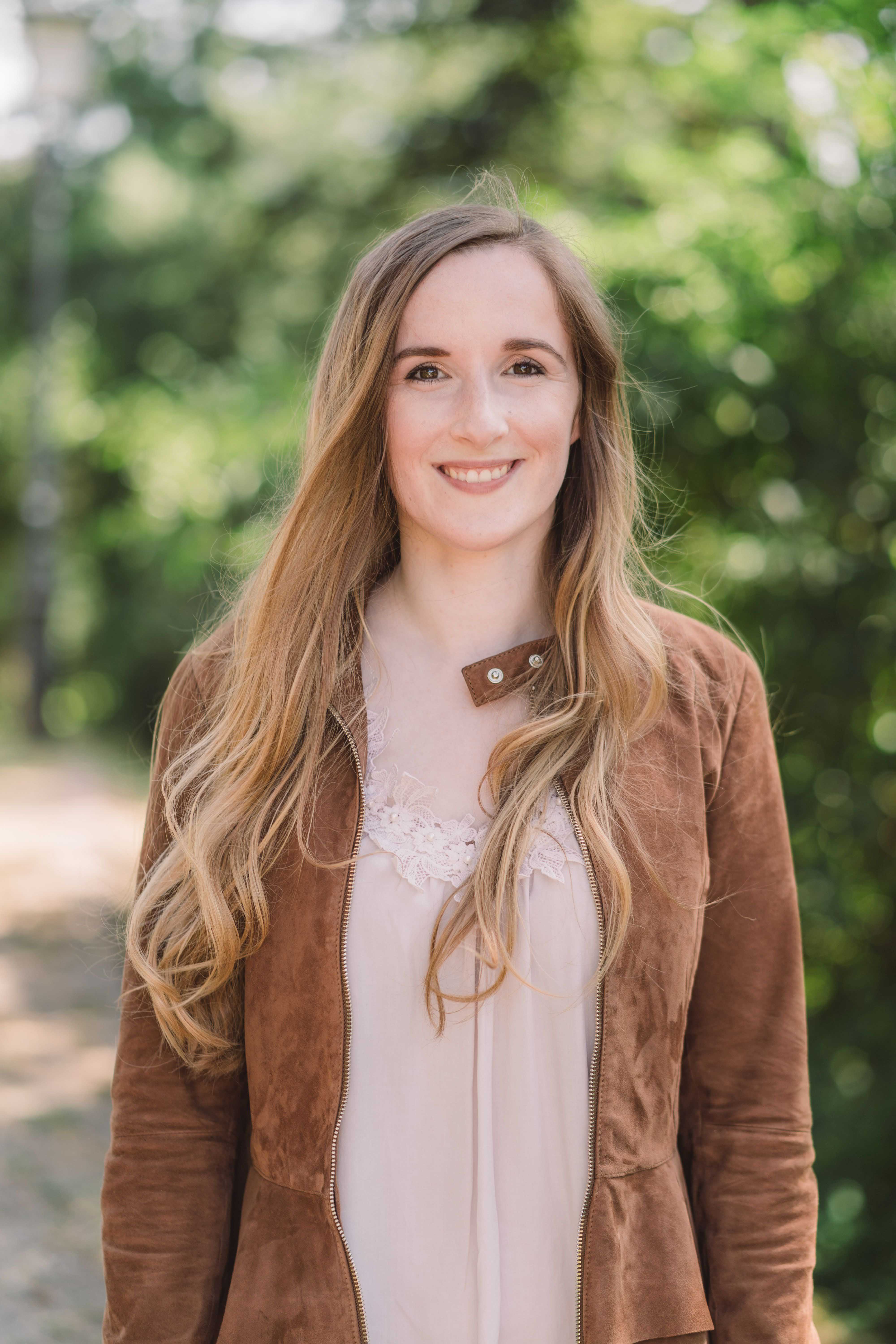 A woman with long, straight hair is seen standing in front of a group of trees, smiling warmly at the camera.