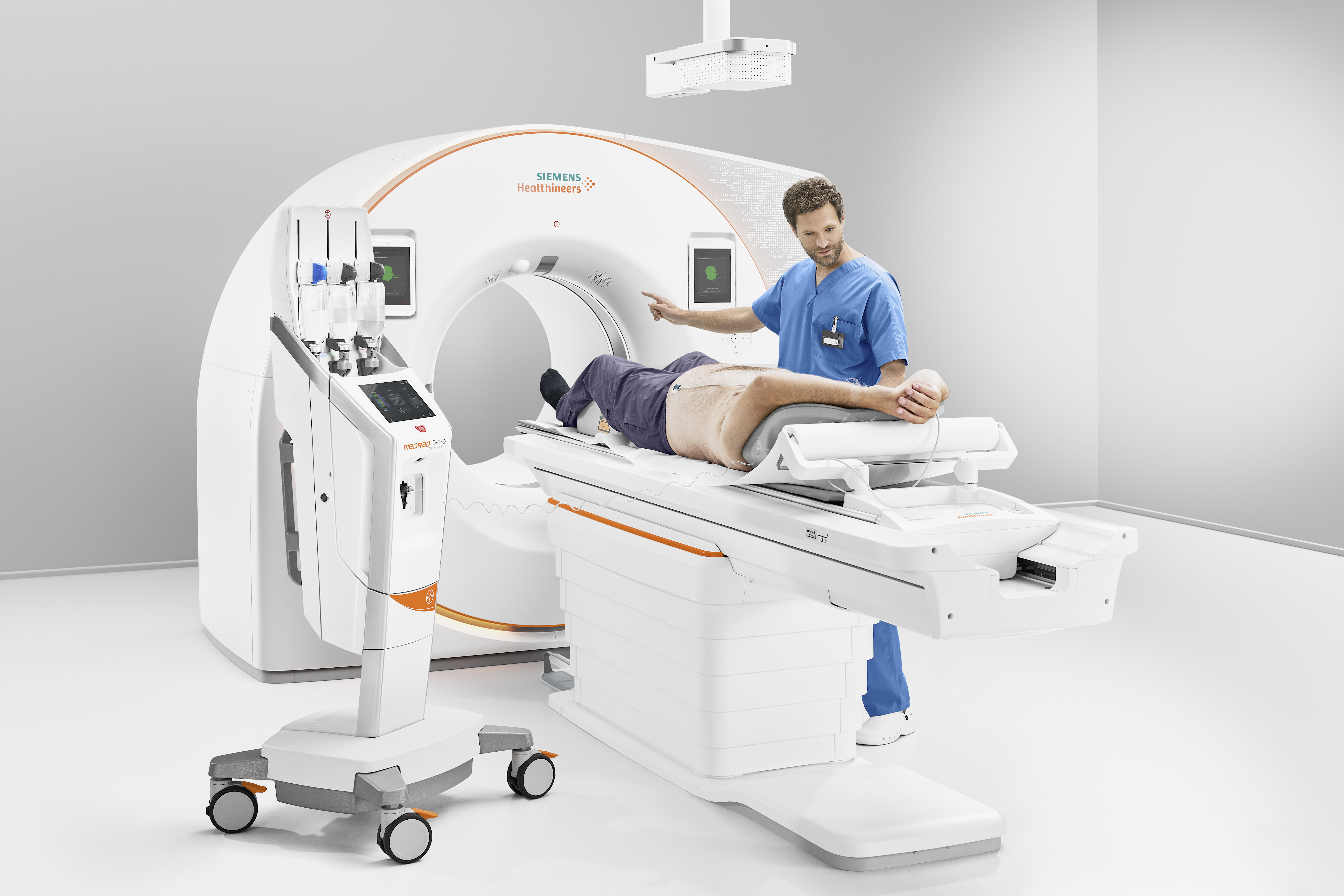 Foto eines PC-CT Geräts mit Ringtunnel, der Strahlenquelle und Detektor enthält, und dem Untersuchungstisch, auf dem eine Person liegt. Ein Arzt erklärt dem Patienten etwas.