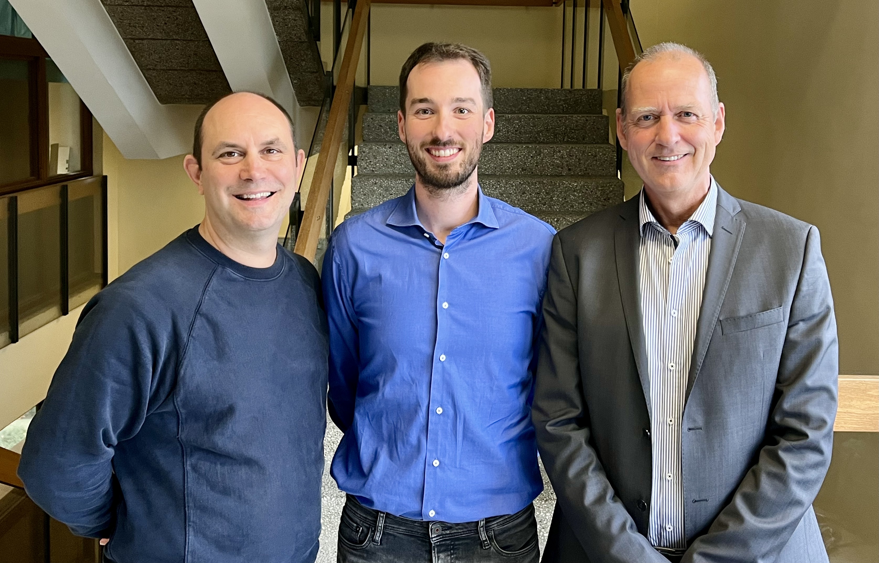 Three men are standing next to each other in a stairwell, smiling friendly into the camera.