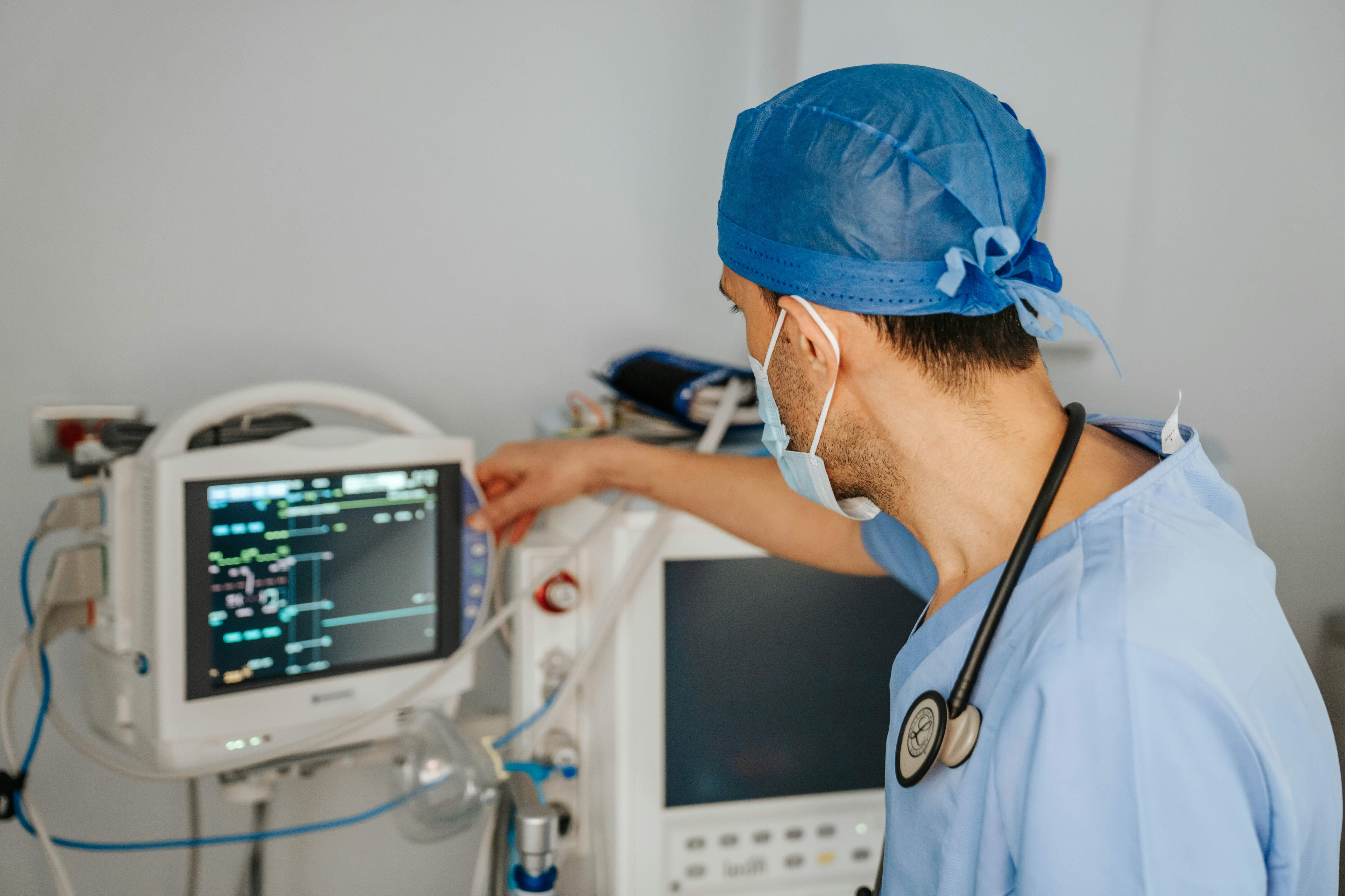 Ein Arzt mit Stethoskop sitzt in OP-Kleidung vor einem Patientenmonitor.