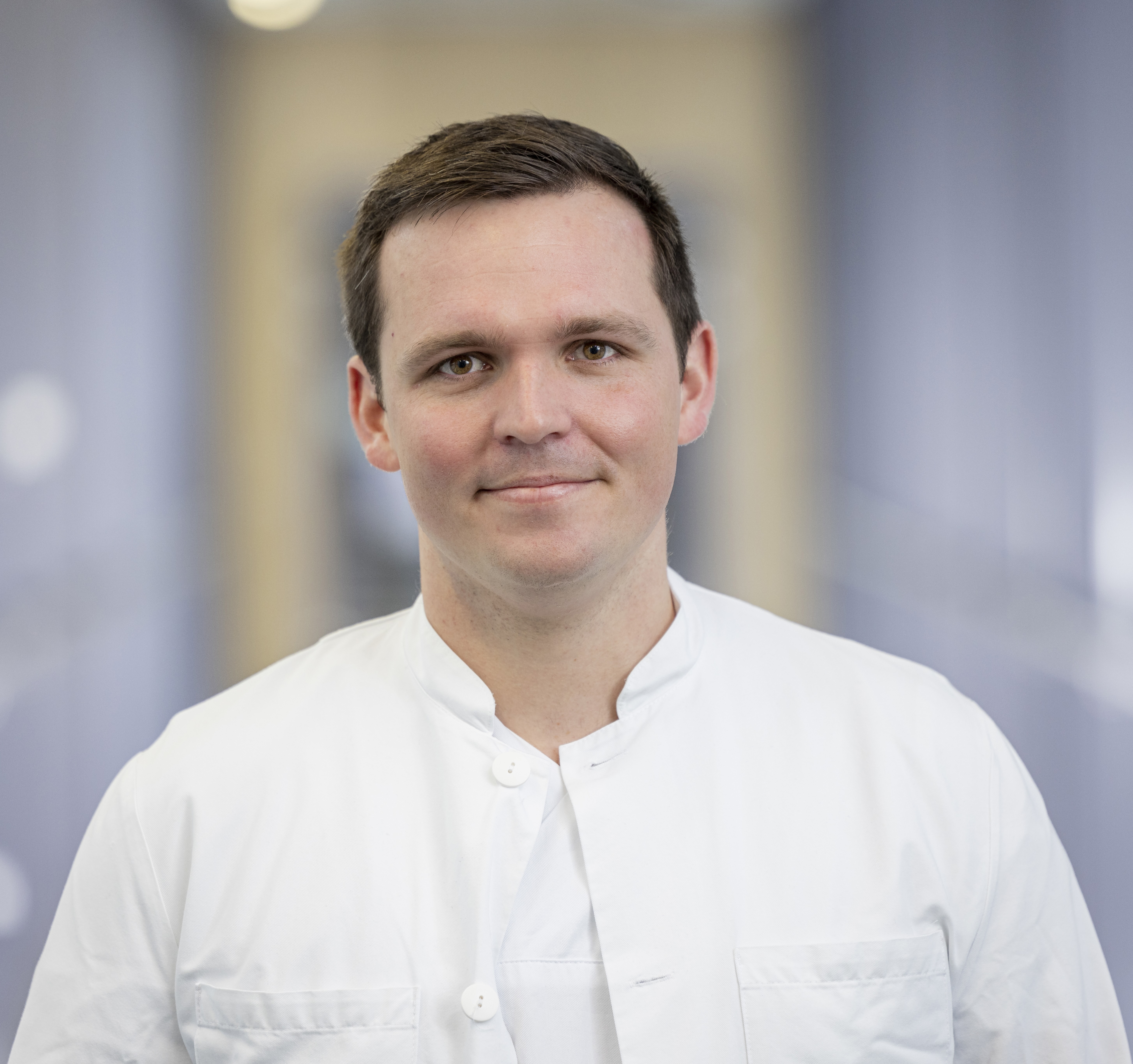 Portrait of a friendly, smiling young man with short, dark hair in a white coat.