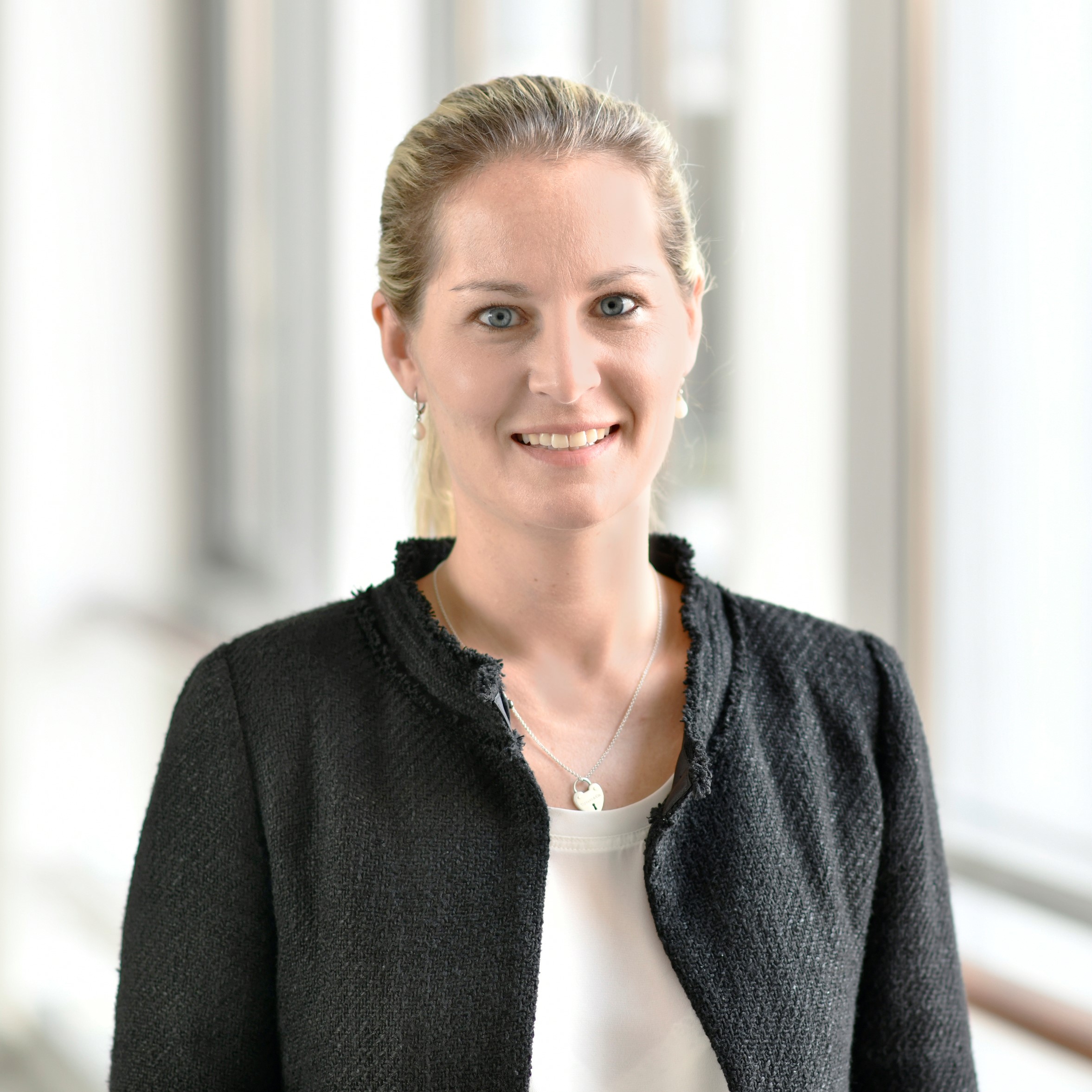 Portrait photo of a friendly smiling blonde woman with tied up hair wearing a black jacket over a white shirt.