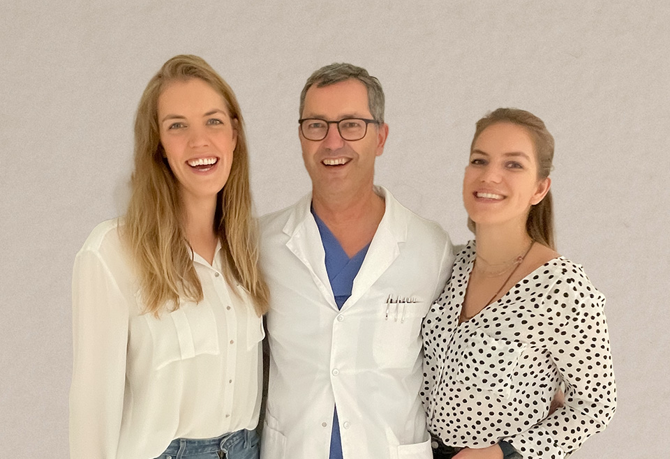 Ein Mann im weißen Arztkittel steht in der Mitte zwischen zwei Frauen.