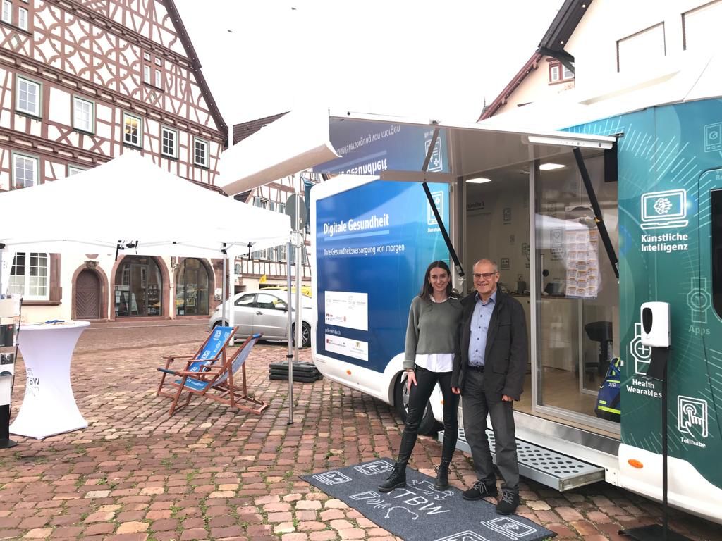 Vor dem Truck stehen zwei Menschen: links eine Frau mit langen braunen Haaren, rechts ein Mann mit dunkler Jacke und blauem Hemd.