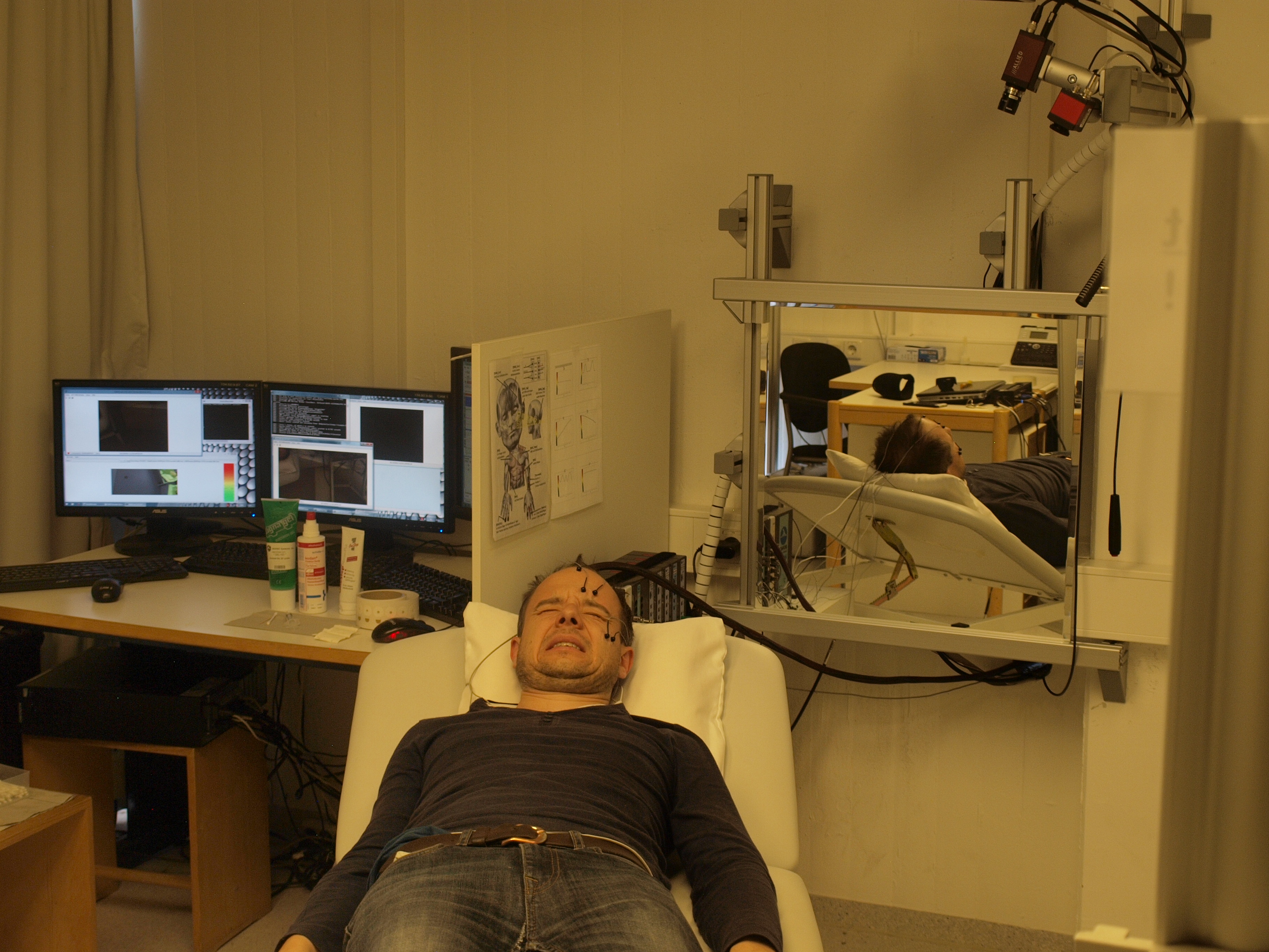 A man lies on a couch with his face contorted in pain. He is connected to a computer system via electrodes.