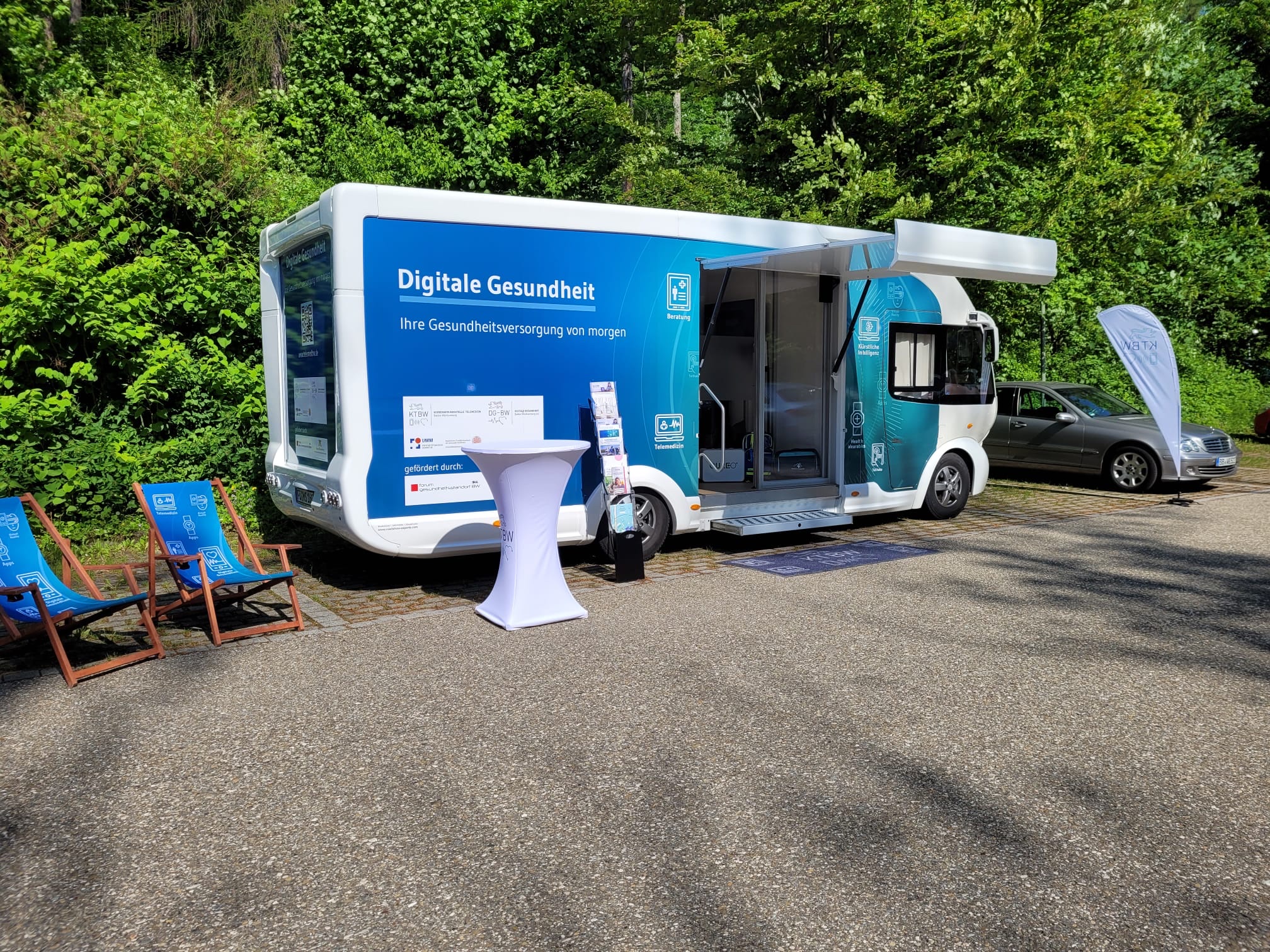 Der Truck steht auf einem Parkplatz in der Sonne vor grünen Bäumen und Büschen.