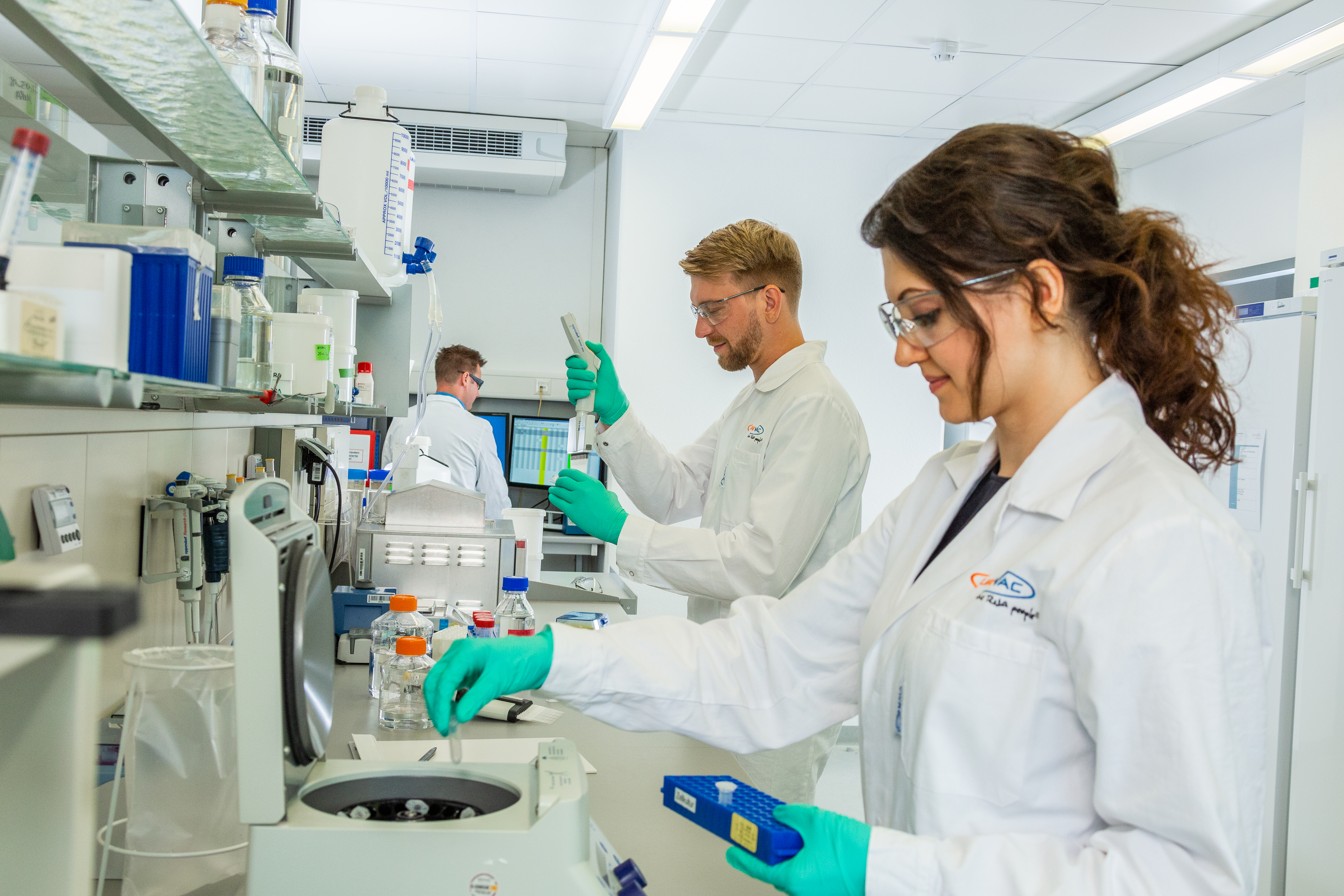 The picture shows two people in a laboratory. They both wear laboratory coats, protective gloves and glasses. While one person is pipetting, the second person is working on a centrifuge.