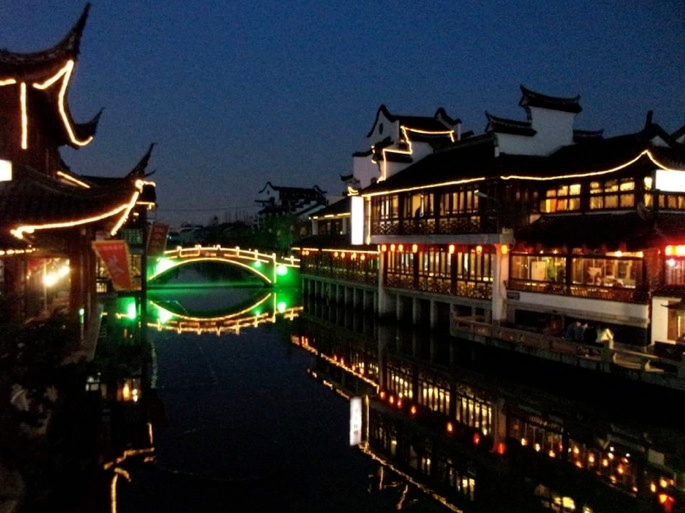 Zu sehen sind hell und festlich beleuchtete Häuser in chinesischem Baustil, die an einem Fluss gelegen sind über welchen eine grün beleuchtete Brücke führt.