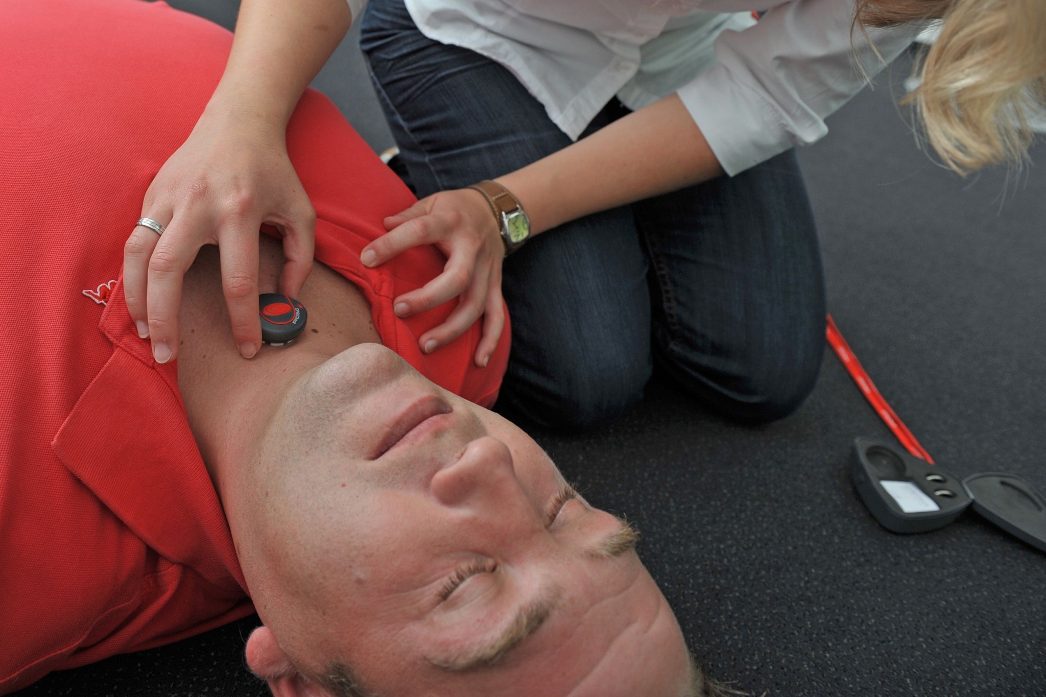 Zu sehen ist ein Mann, der bewusstlos auf dem Boden liegt und von einer weiteren Person den neuen Erste-Hilfe-Sensor an den Kehlkopf gedrückt bekommt.