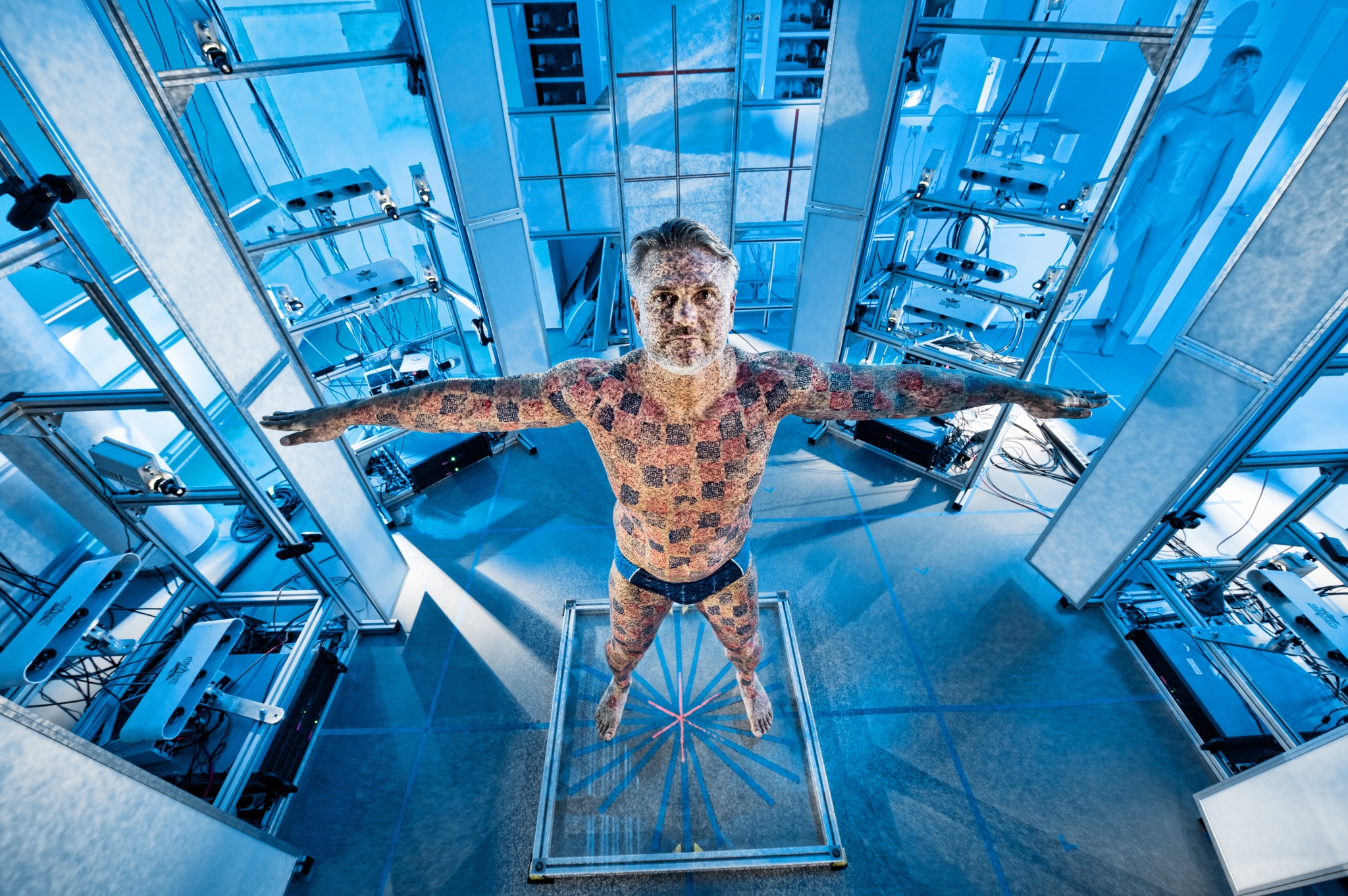 Ein Mann, der im Labor im Ganzkörperscanner steht, und dessen Körper von blauen Vierecken beleuchtet wird.
