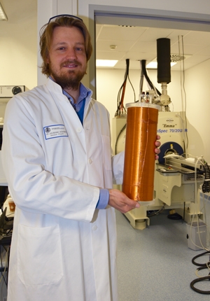 Zu sehen ist der Forscher Dr. Jan-Bernd Hövener mit einer armlangen Magnetspule in der Hand. Im Hintergrund sieht man einen großen Magnetresonanztomografen.
