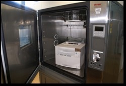 The photo shows a metal cupboard with a white laser printer inside. The door of the cupboard is open.