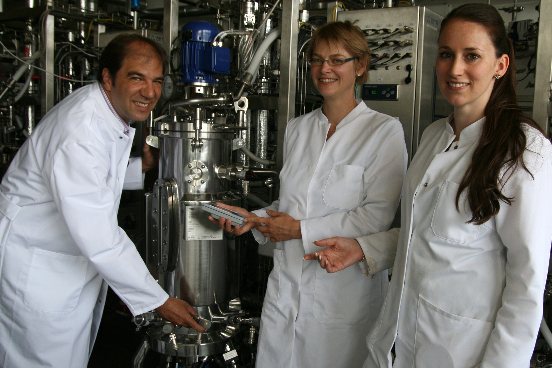 A man and two young women standing next to a bioreactor and smiling into the camera.