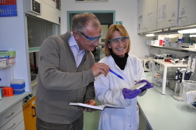 Prof. Dr. Martin Elmlinger and his colleague Marion Eisenhauer in the laboratory. Elmlinger is holding a ball-pen in his hand, pointing at a microtitre plate Marion Eisenhauer is holding in her hand.