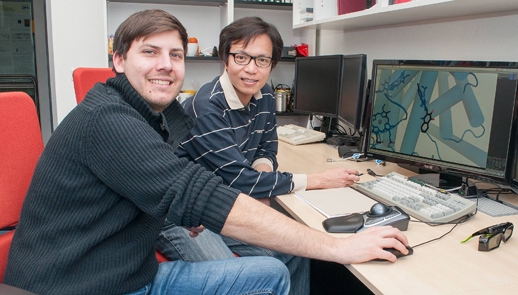 The photo shows two young men sitting in front of a computer graphic station. Both are smiling into the camera.
