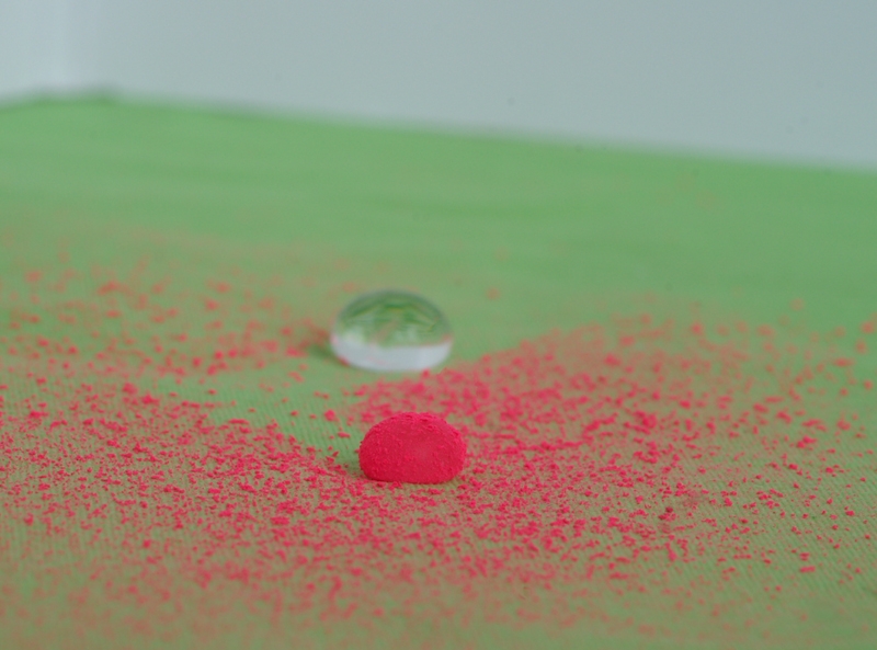 Stark vergrößerte Stoffoberfläche mit roten Partikeln, die aufgrund des Lotuseffekts von einem Wassertropfen aufgesammelt werden.