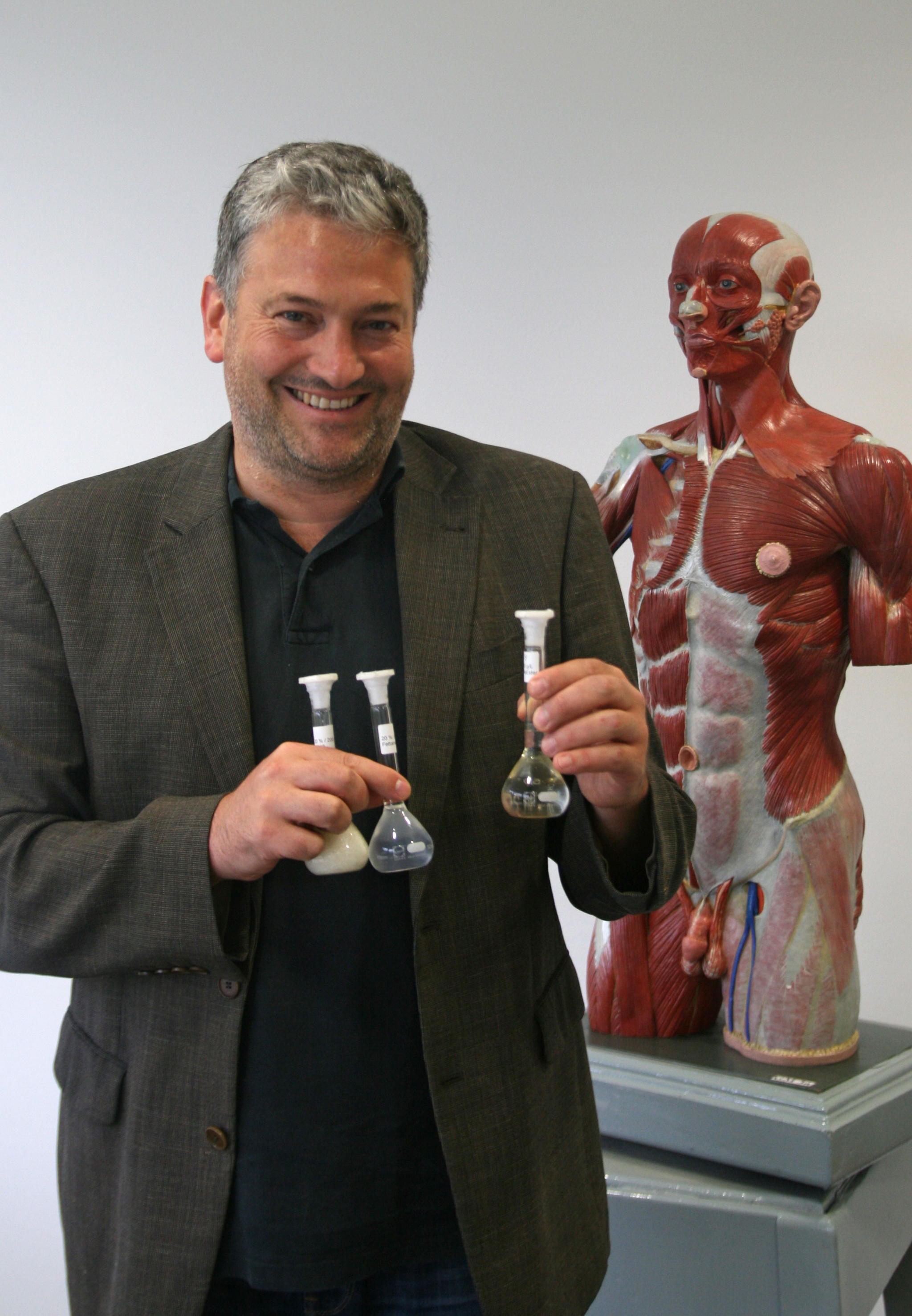 Bernhard Hirt, stehend vor einem anatomischen Modell eines Menschen, hält drei Glaskolben in den Händen.