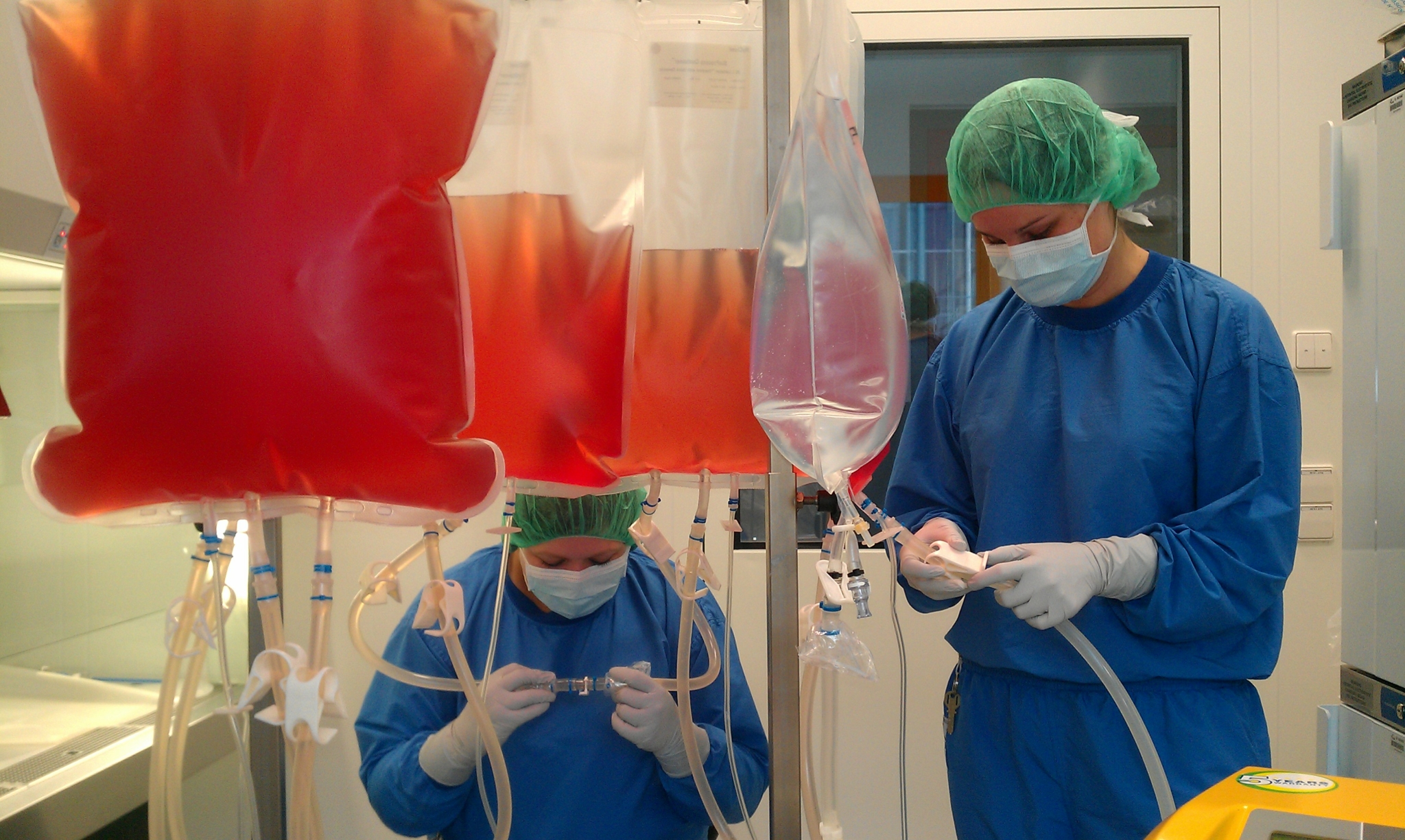 Two people in blue sterile clothing, wearing a face mask and a green hair cover, handling plastic tubes filtering the antibody solution under sterile conditions. Bags containing a red/transparent liquid are shown in the foreground.