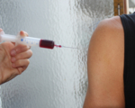 Photo of a hand holding a syringe containing a dark fluid towards a naked upper arm.