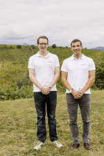 Zwei Männer stehen nebeneinander auf einer Wiese, die sich im Hintergrund mit Büschen fortsetzen. Sie tragen weiße Polohemden mit Unternehmenslogo und blaue Jeans.