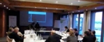 IN einem Raim sitzen Leute um einen Tisch und schauen sich eine Präsentation an.