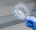 Hand holding a round plastic disk with numerous channels and chambers.