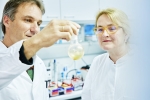 Die beiden Forschenden in Laborkitteln im mikrobiologischen Labor, einen Glaskolben mit beigefarbenem Epifadin betrachtend.
