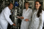 Ein Mann und zwei junge Frauen stehen neben einem Bioreaktor und lächeln in die Kamera.