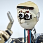 Close-up photo of robot with a human-like head, holding up a hand with fingers.