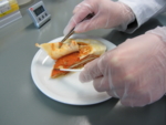 Zu sehen sind zwei Hände in Gummihandschuhen, die ein Fertiggericht mit Pinzette und Skalpell untersuchen.
